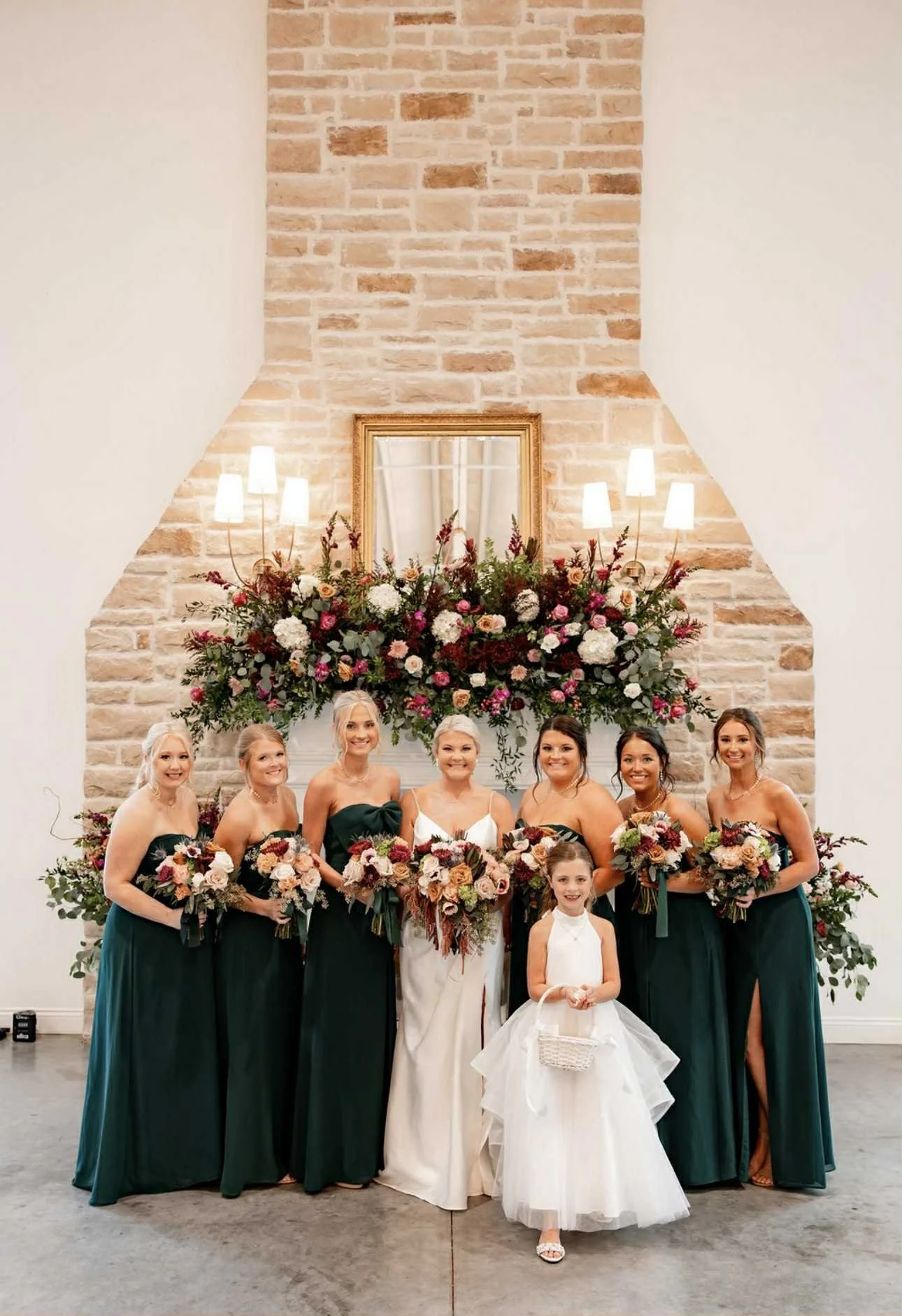 Group of women in formal dresses holding bouquets, standing in front of a large floral arrangement and mirror on a brick wall, at a wedding event.
