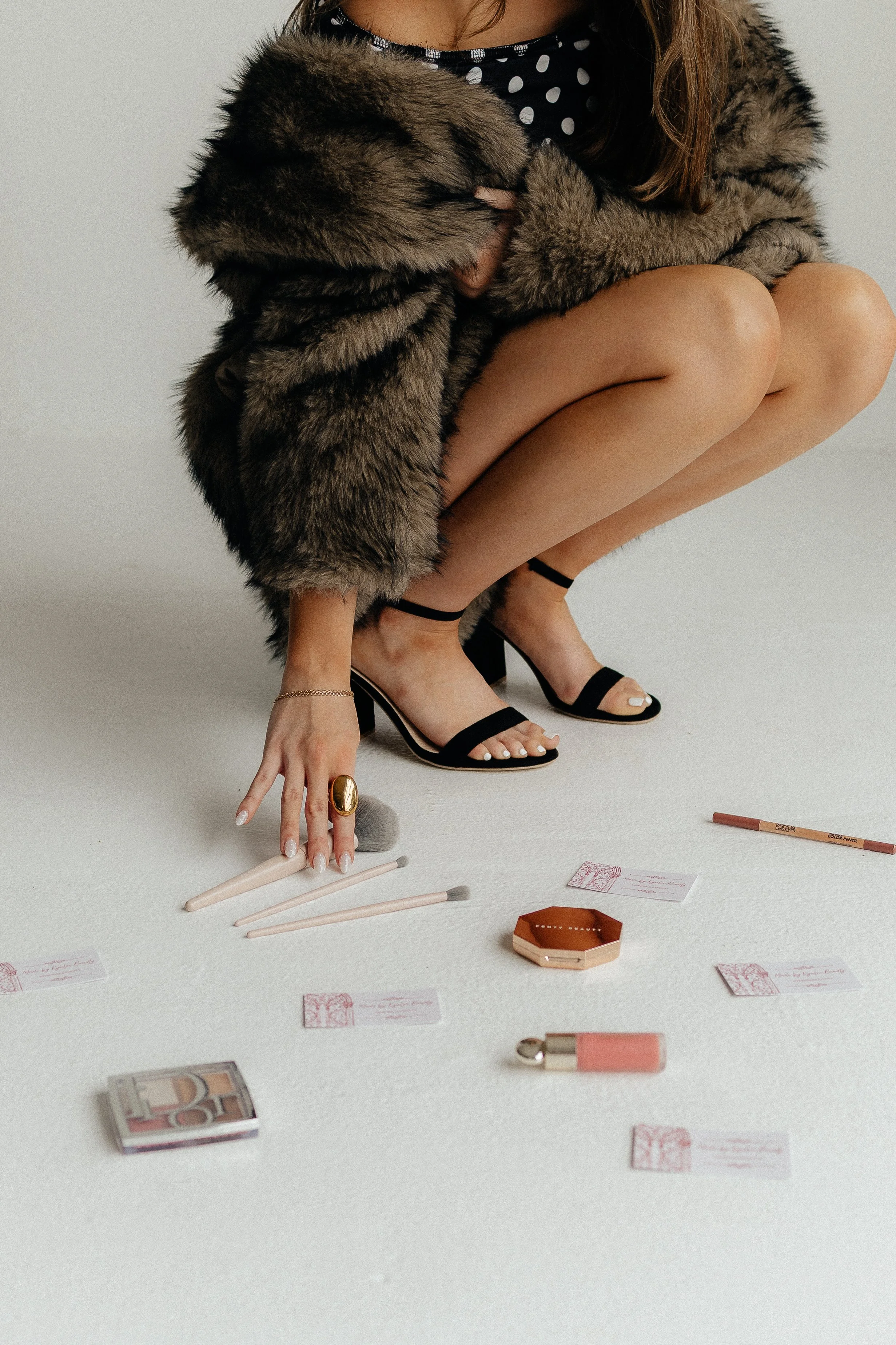 A woman wearing a polka dot top and black high heels with crossed legs, a fur coat, and jewelry, crouching on a white floor surrounded by makeup and beauty products, including brushes, lipstick, and small cards.