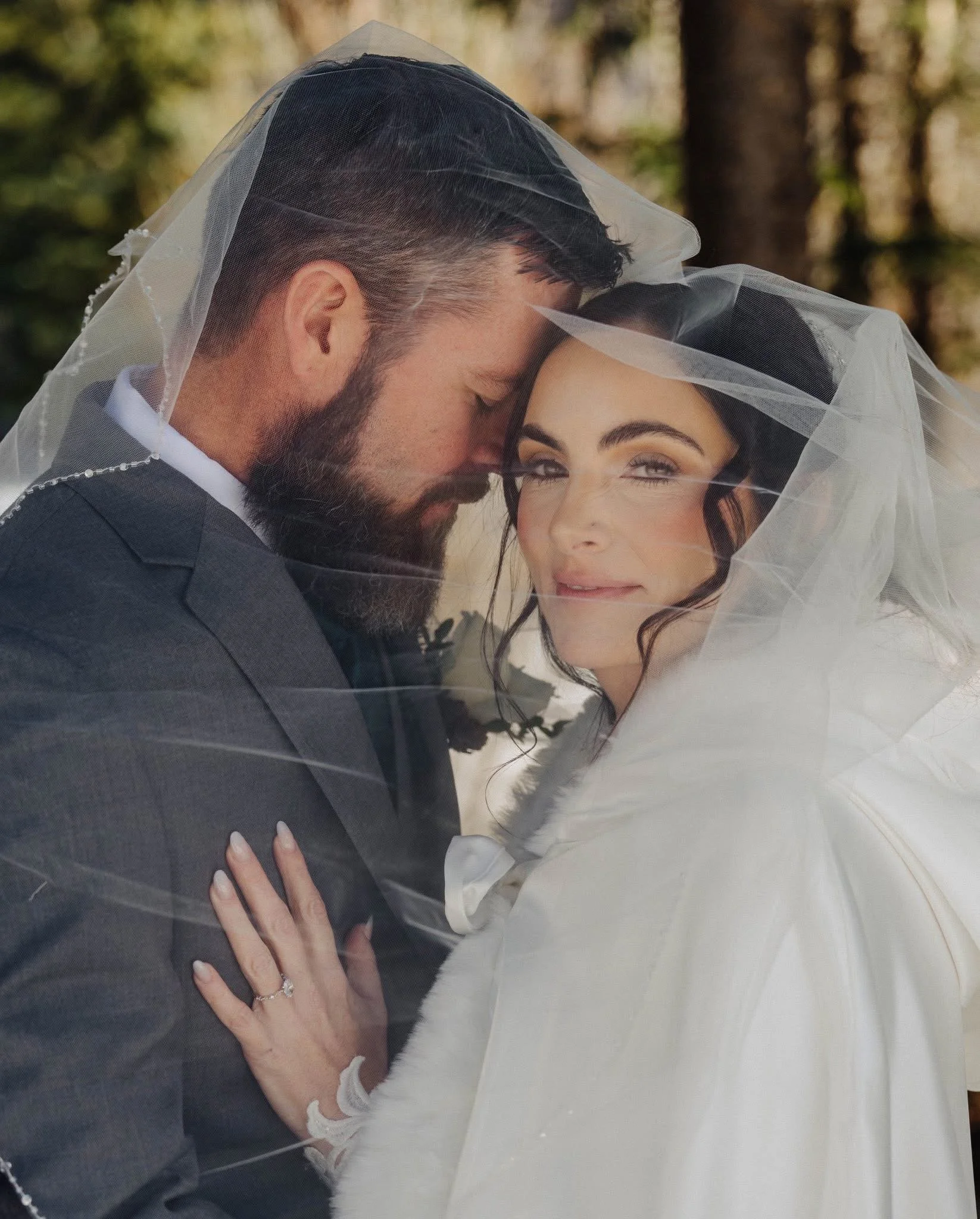 A bride and groom embrace under a sheer white bridal veil outdoors during daytime. The groom has a beard and mustache, and wears a dark suit. The bride has dark hair, styled with waves, and wears a white dress. Her hand, with rings, is resting on the groom's chest.