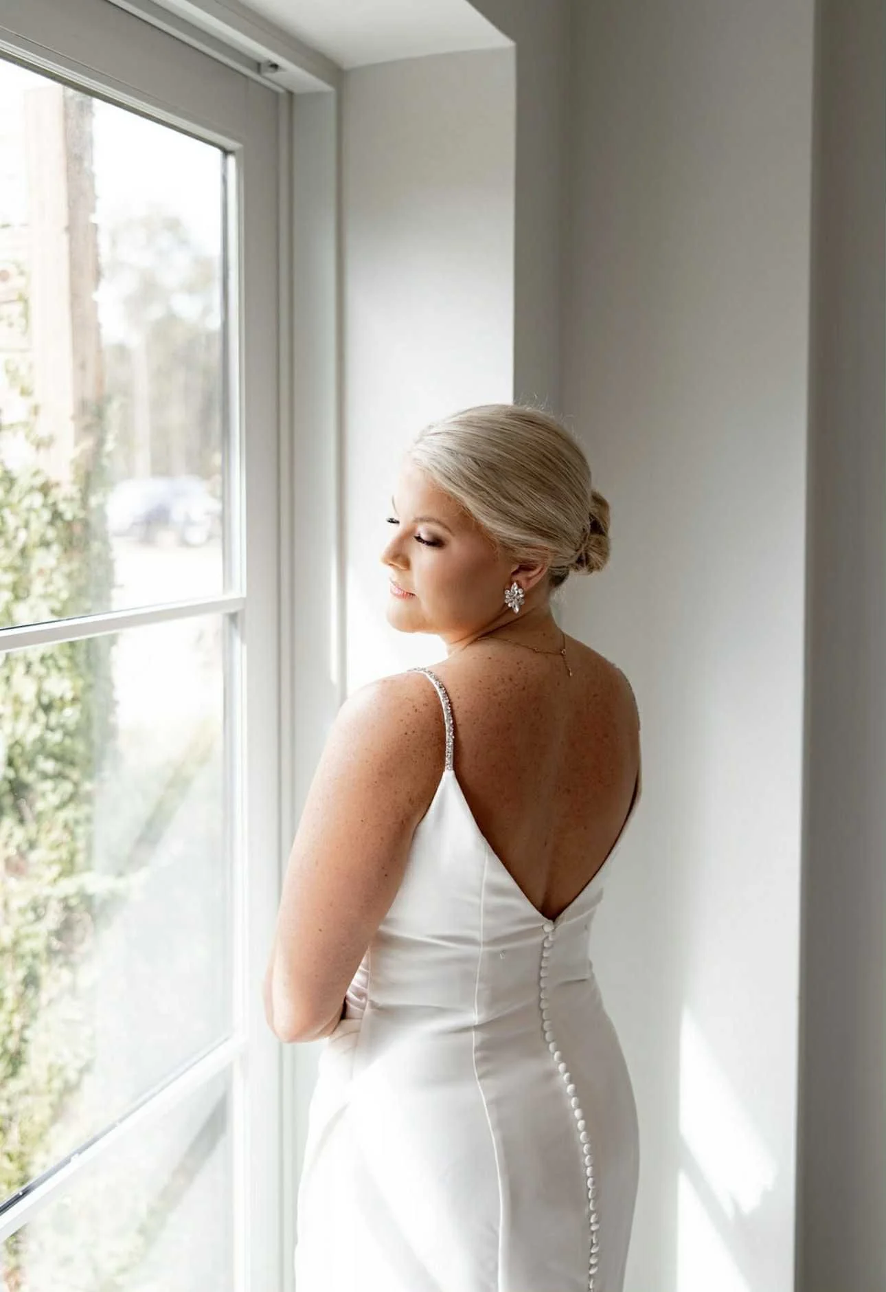 A bride with blonde hair styled in an elegant updo standing by a window, wearing a white wedding gown with buttons on the back and floral earrings.