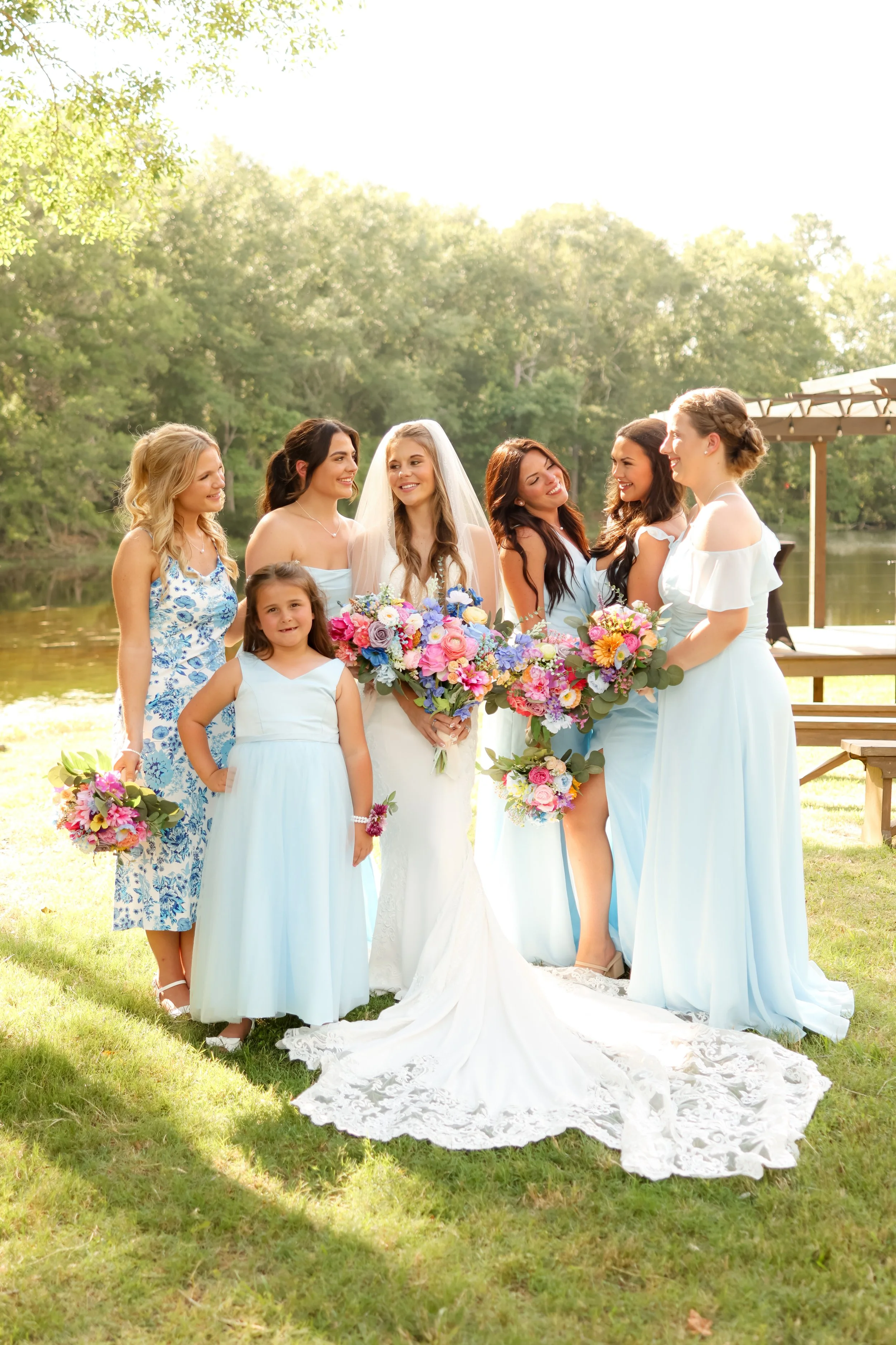 A bride and six women, likely bridesmaids, standing outdoors by a lake surrounded by trees. They are dressed in light blue and white dresses, holding colorful bouquets, smiling and engaging with each other on a sunny day.