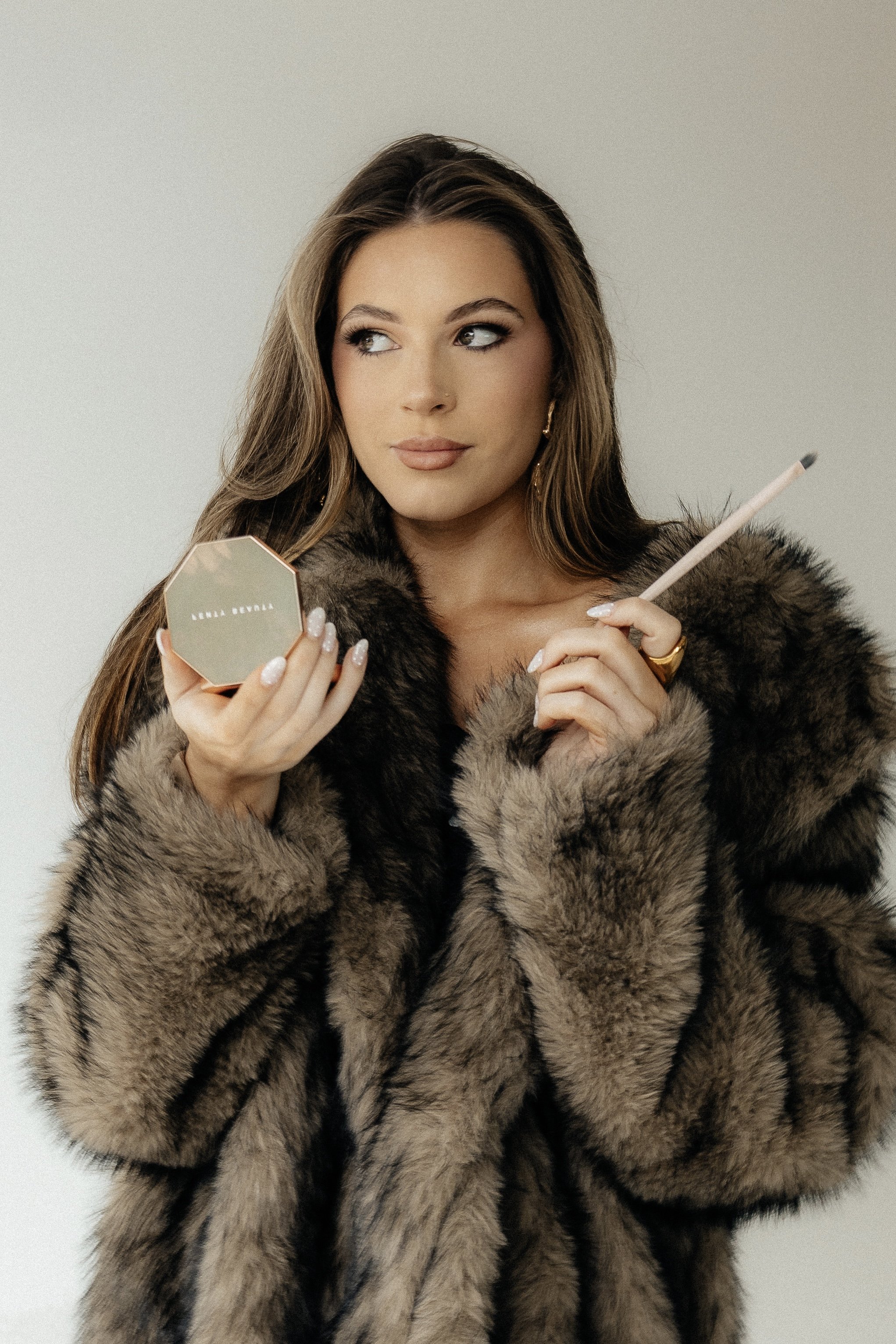 Woman with long brown hair wearing a fur coat applying makeup using a small mirror and a makeup brush.