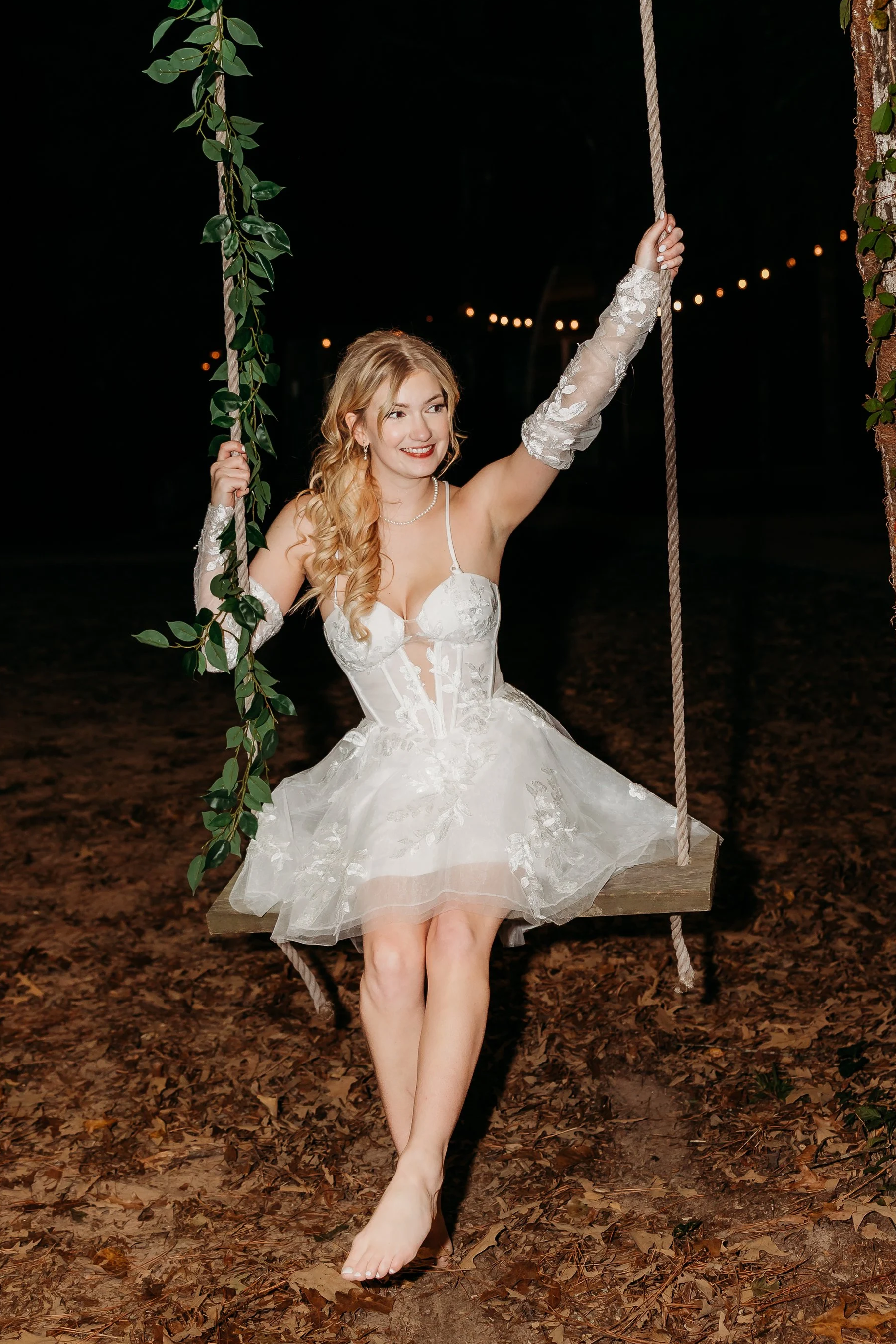 A woman in a white lace dress sitting on a wooden swing with rope, surrounded by leaves and fallen brown leaves on the ground at night