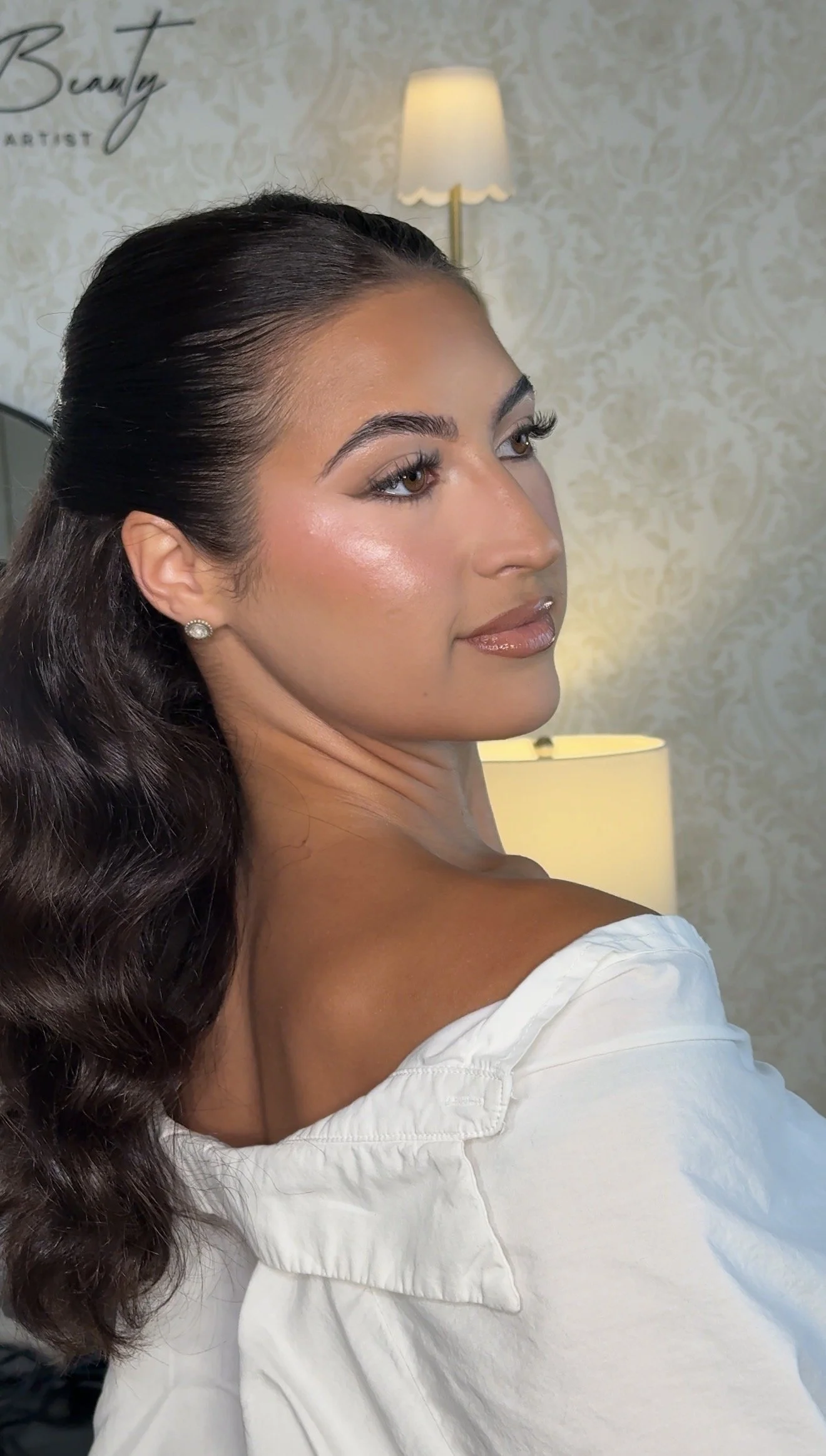 Portrait of a woman with long wavy dark hair, wearing pearl earrings and a white off-shoulder top, in a warmly lit room with patterned wallpaper and a lamp in the background.