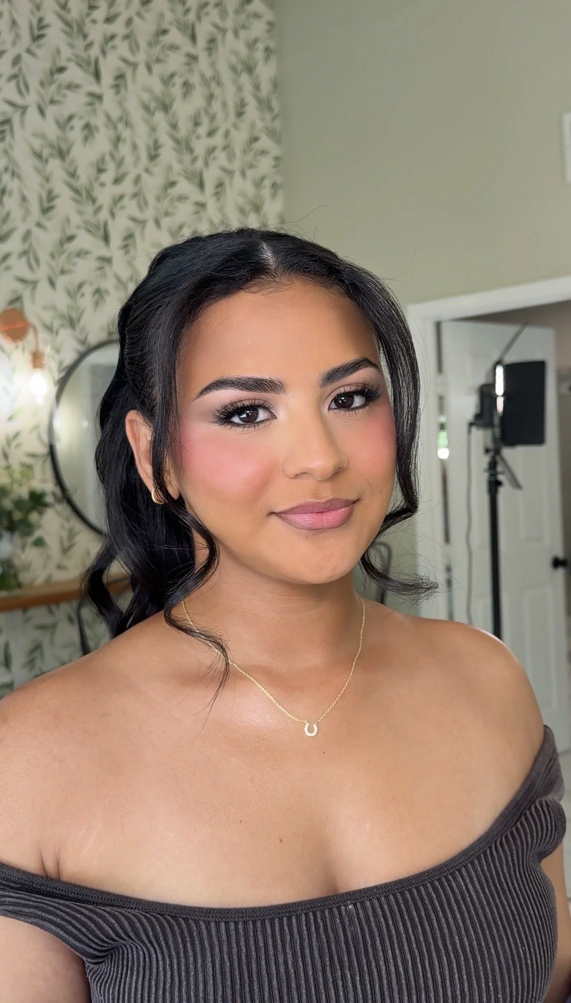A close-up of a woman with dark, wavy hair, wearing makeup, a necklace, and a off-shoulder dark ribbed top, in a room with green patterned wallpaper and studio equipment in the background.
