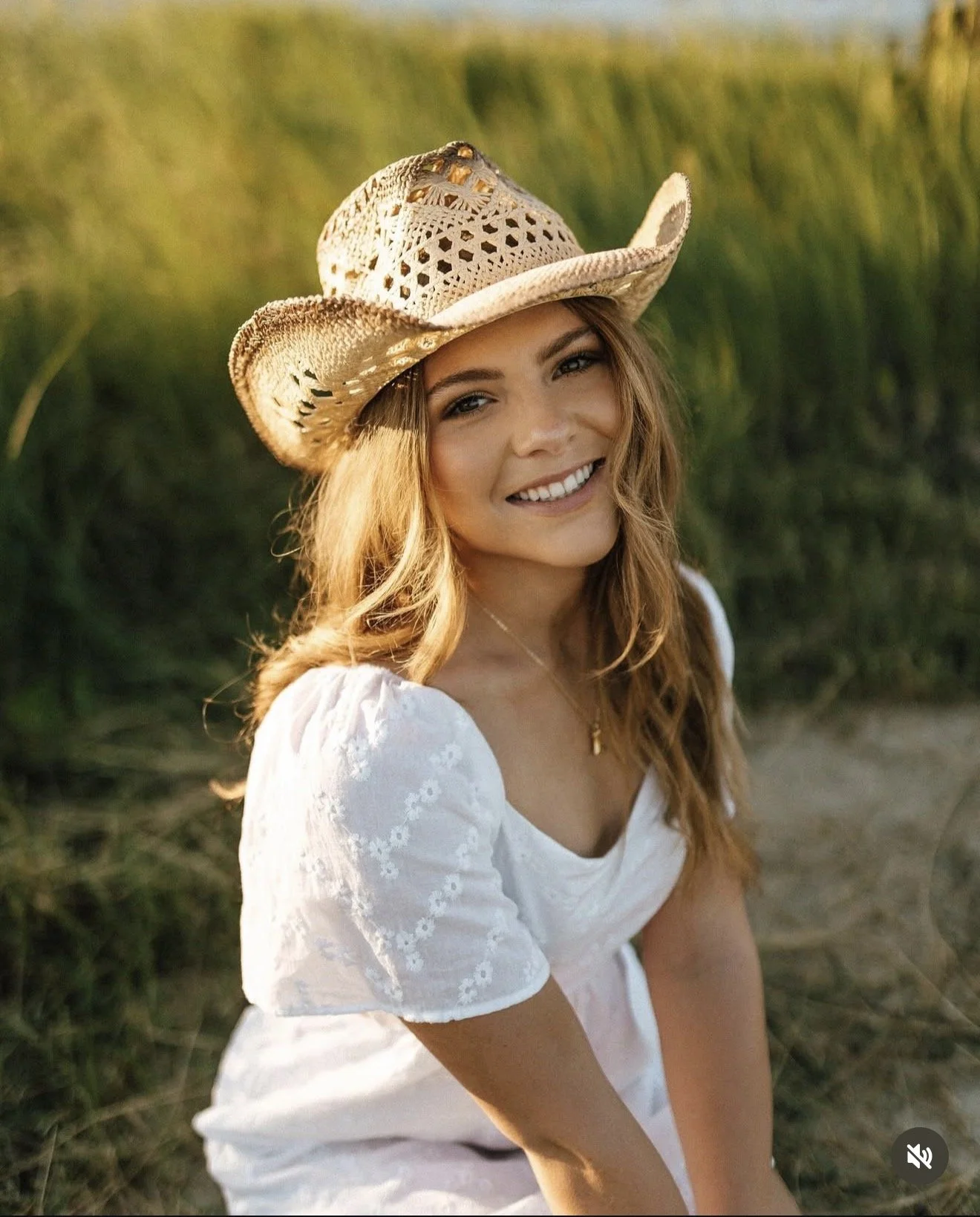 A smiling woman with long, wavy hair wearing a wide-brimmed straw hat and a white dress, sitting outdoors in a grassy area.