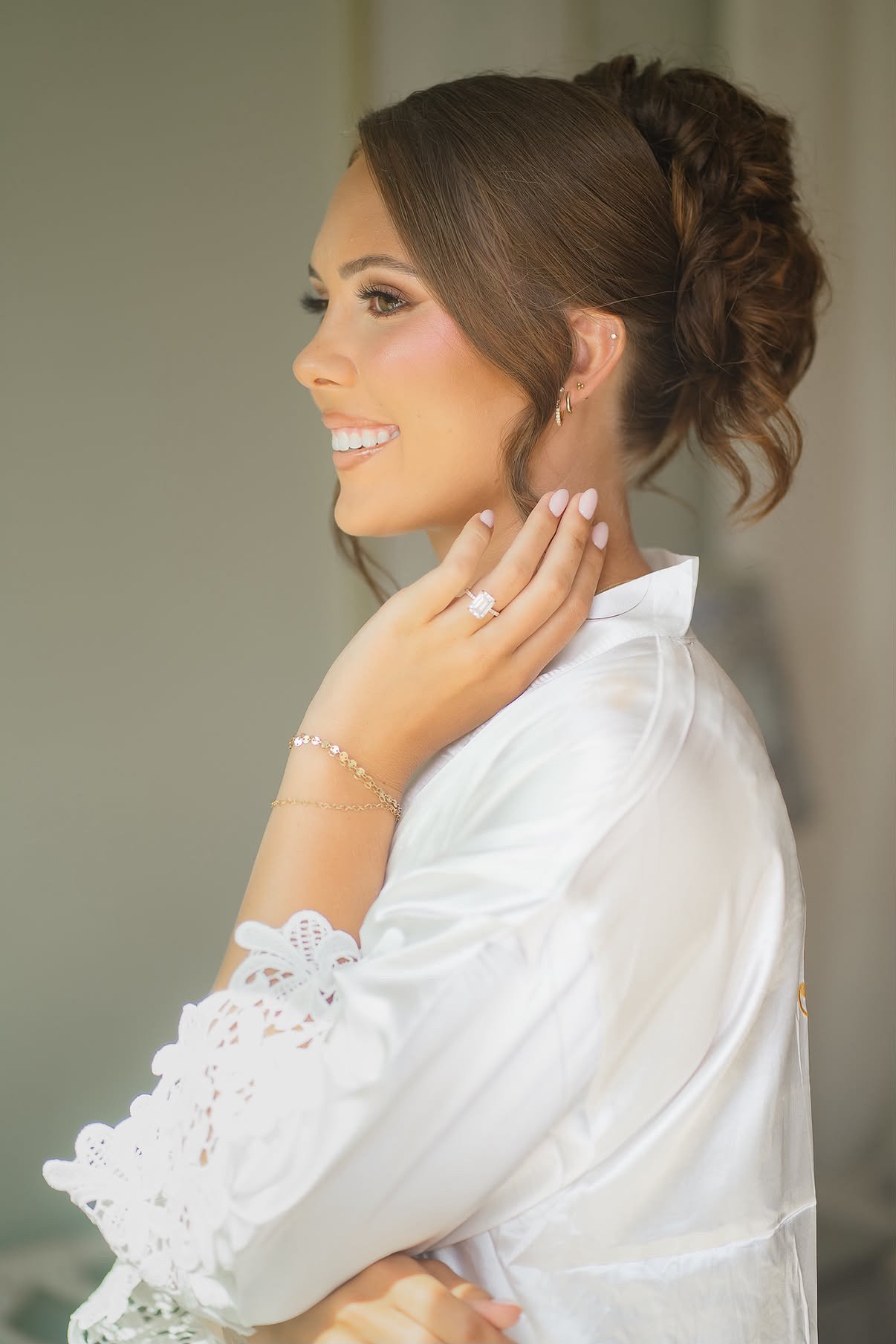 A woman with brown hair styled in an updo, smiling softly, wearing a white satin robe with lace details, a large ring, earrings, a bracelet, and a delicate necklace, looking to her left with her hand resting on her neck.