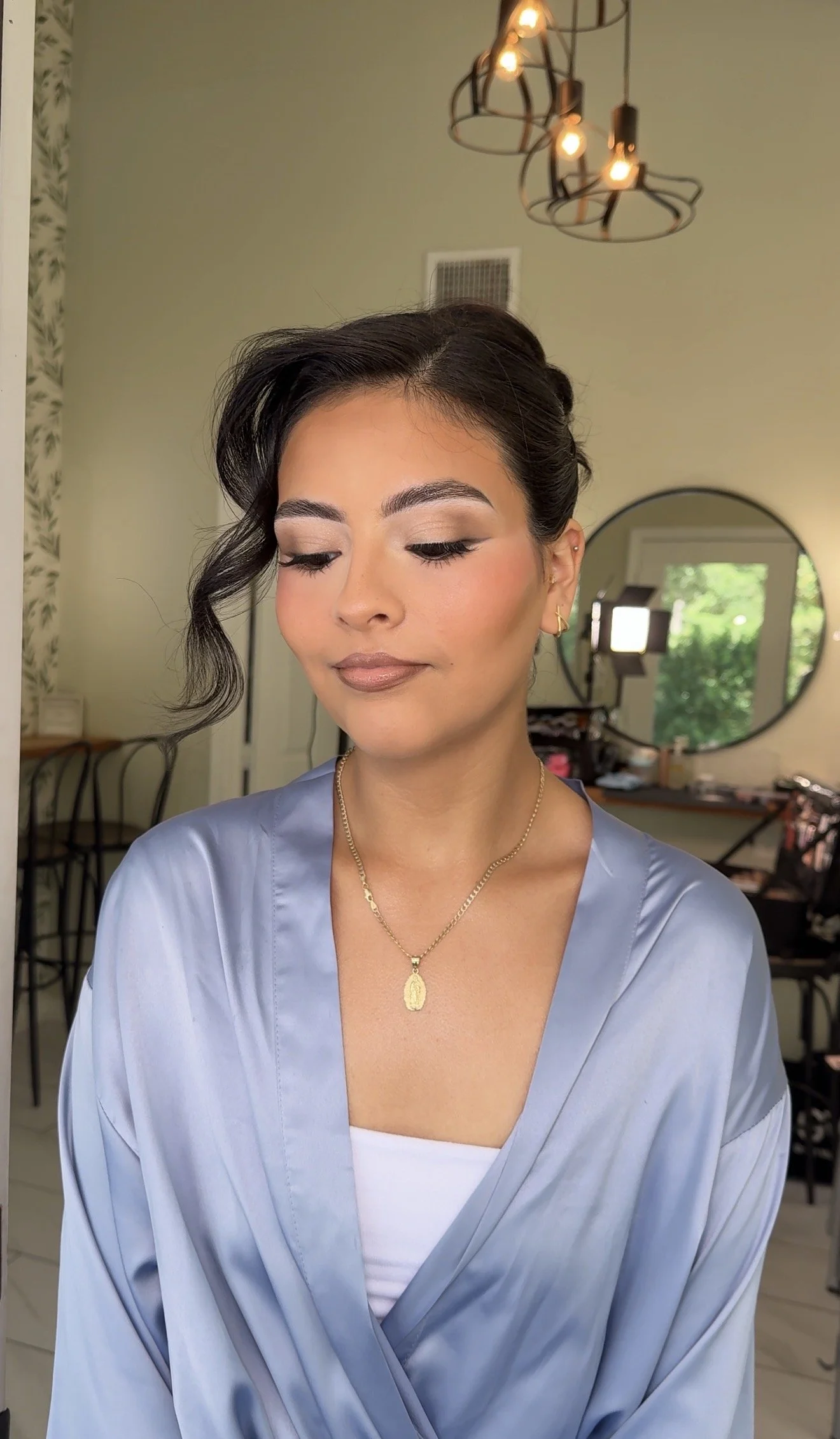 A woman with styled short dark hair, wearing a light blue satin robe, a white undershirt, gold jewelry, and makeup, sitting in a room with a mirror, chairs, and bright lighting fixtures.