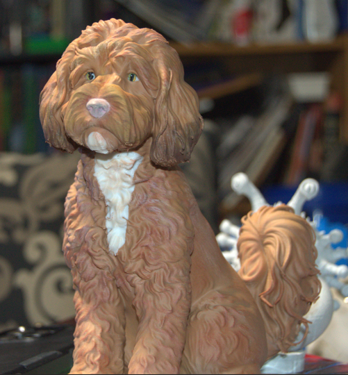 A figurine of a brown and white dog with a curly coat, sitting on a surface with various objects and blurred shelves in the background.