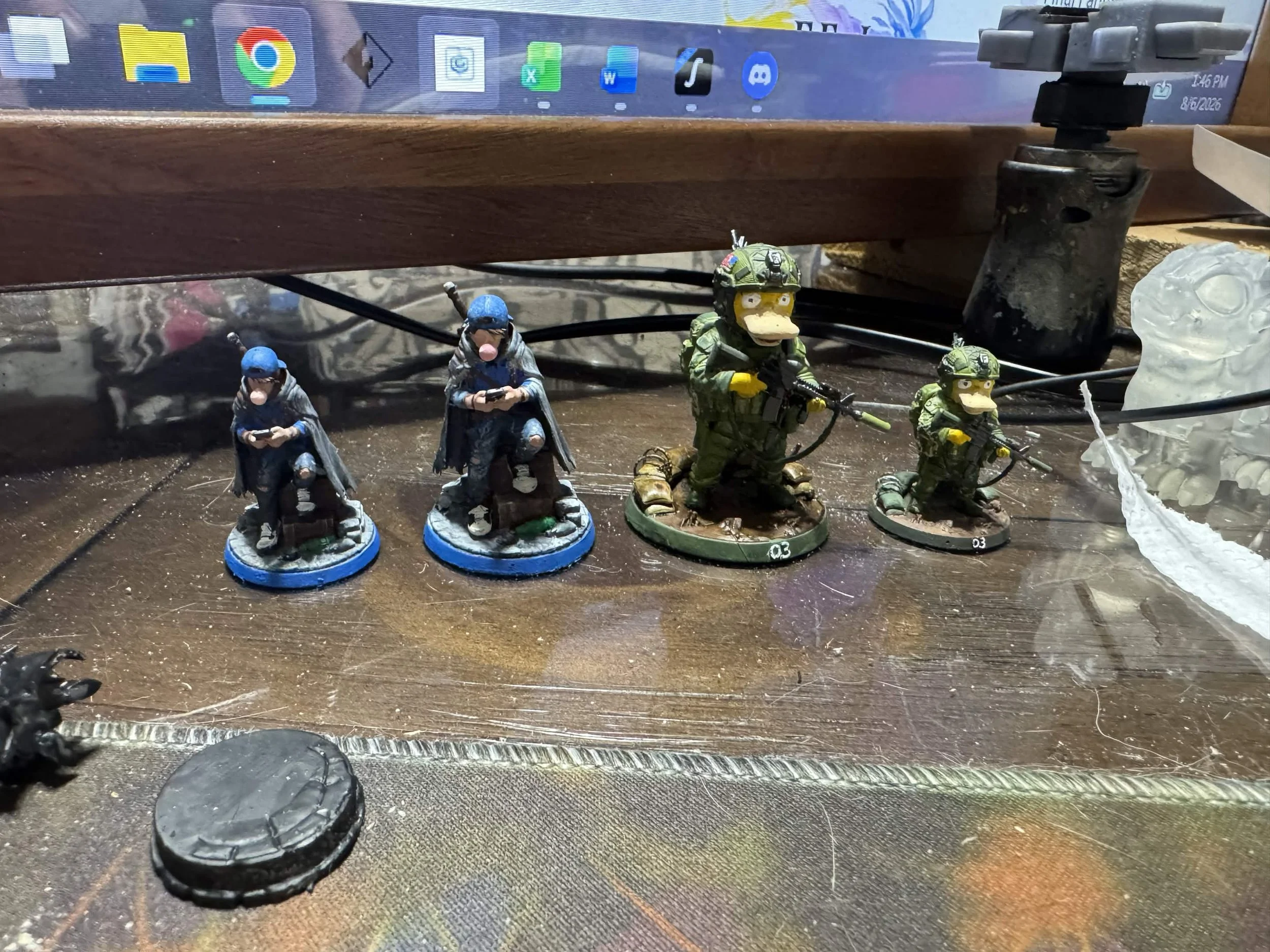Four small figurines of soldiers and a monkey, with a dark desk surface, a computer monitor with icons, a black stand, a plastic bag, and some black wires in the background.