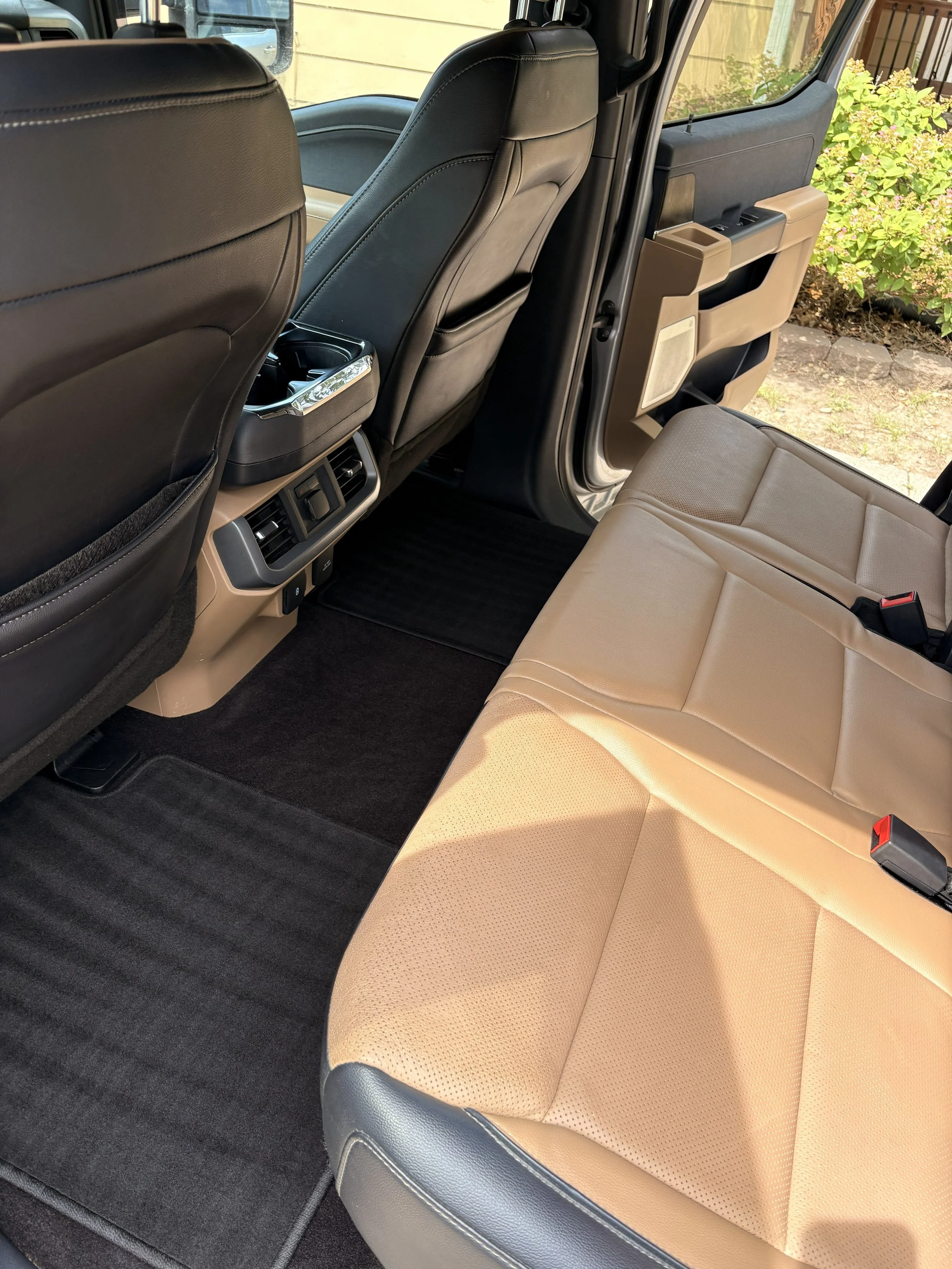 The interior of a vehicle showing beige and black leather seats, rear air vents, and rear seat with a seatbelt buckle visible on the right.