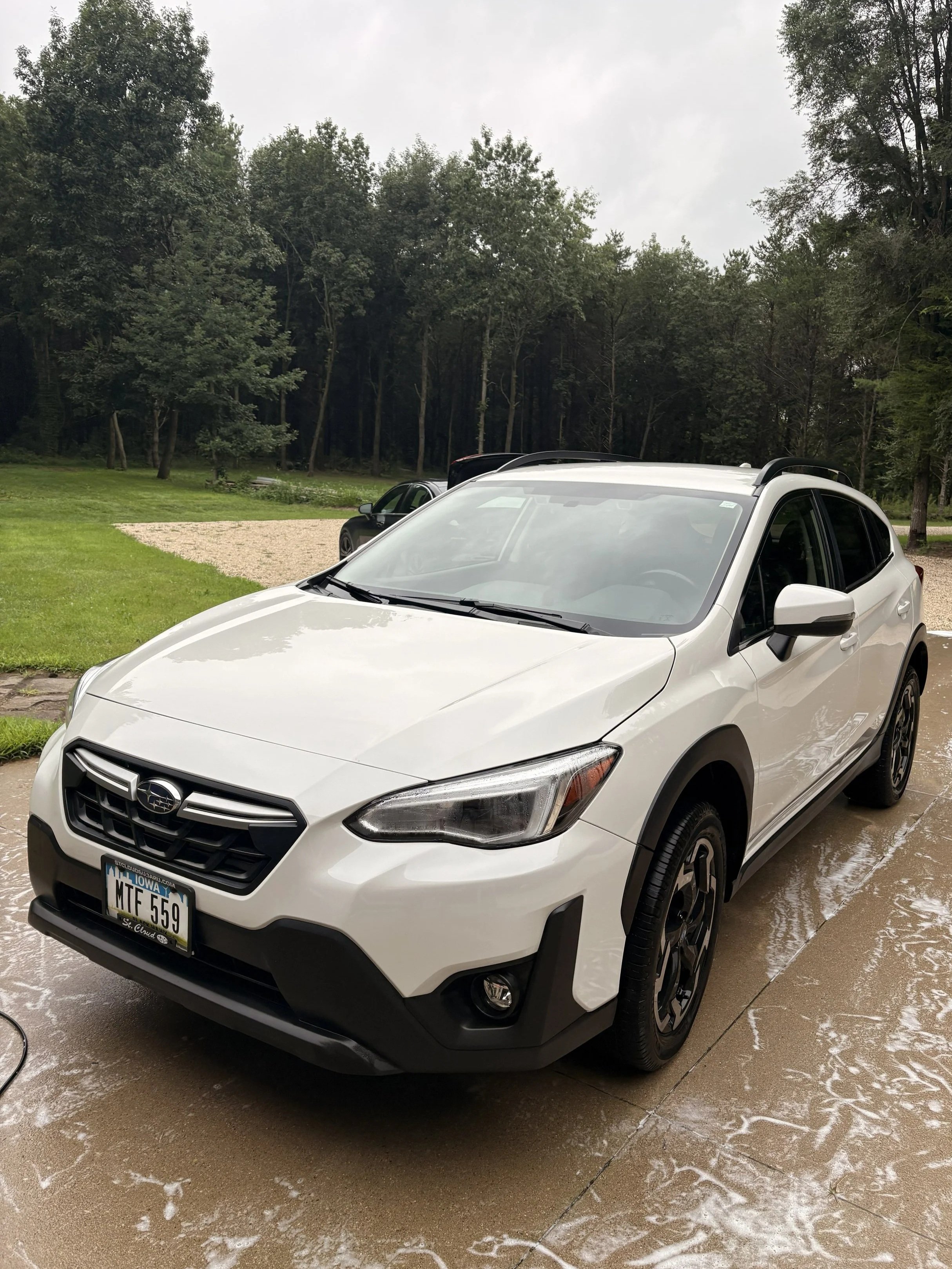 A white Subaru crossover parked on a wet driveway with soap suds, with a green lawn and densely wooded area in the background, under a cloudy sky.