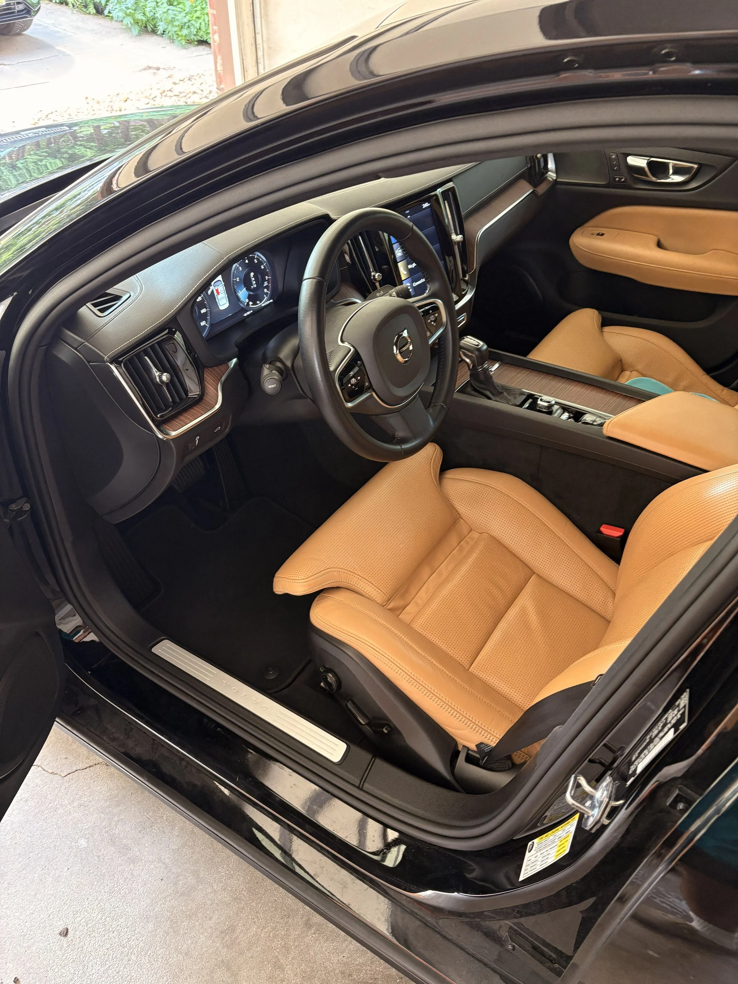 Inside view of a black car with tan leather seats, dashboard, steering wheel, and center console. The door is open, showing the interior layout.
