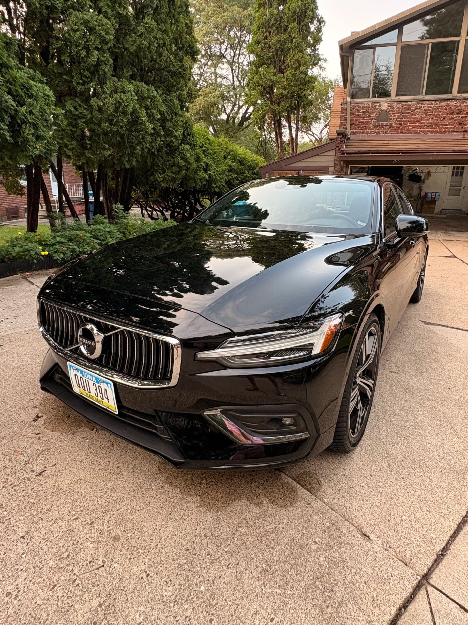 A black Volvo sedan parked on a driveway in front of a house. The driveway is concrete, and there are trees and green bushes nearby. The house has a brick and wood exterior with a garage door open, showing some items inside.