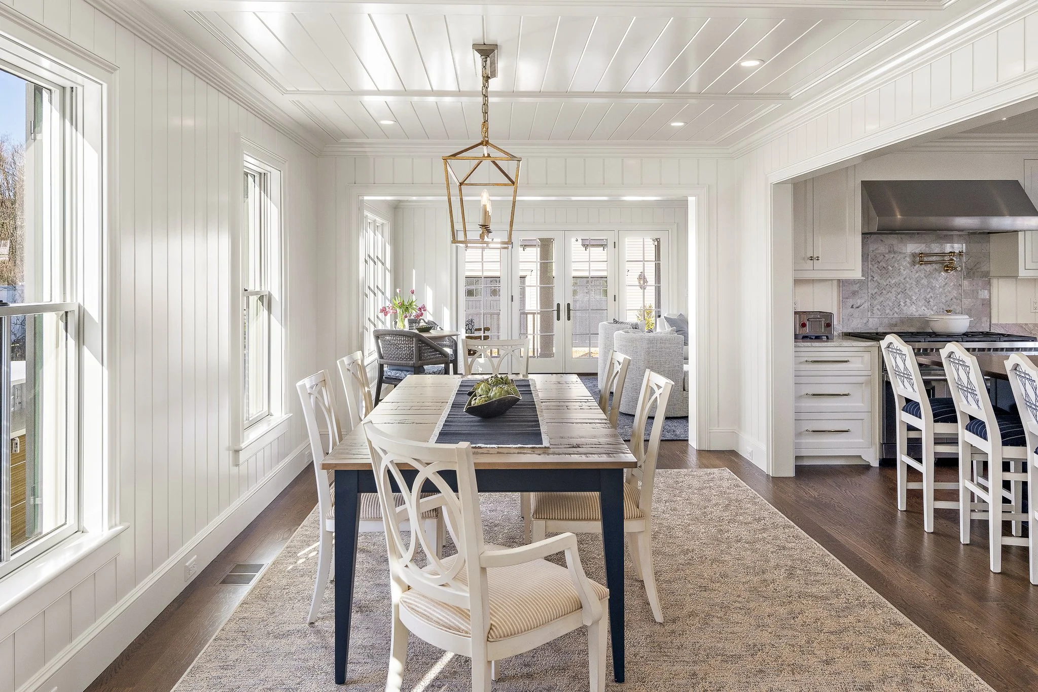 Bright dining area with large windows, white paneled walls, and a wooden table surrounded by white chairs. A modern gold chandelier hangs over the table, which has a black runner and a fruit bowl. In the background, there are glass doors leading outside, and a cozy sitting area with chairs and a sofa. The kitchen on the right has white cabinetry, marble backsplash, and a stainless steel range hood.