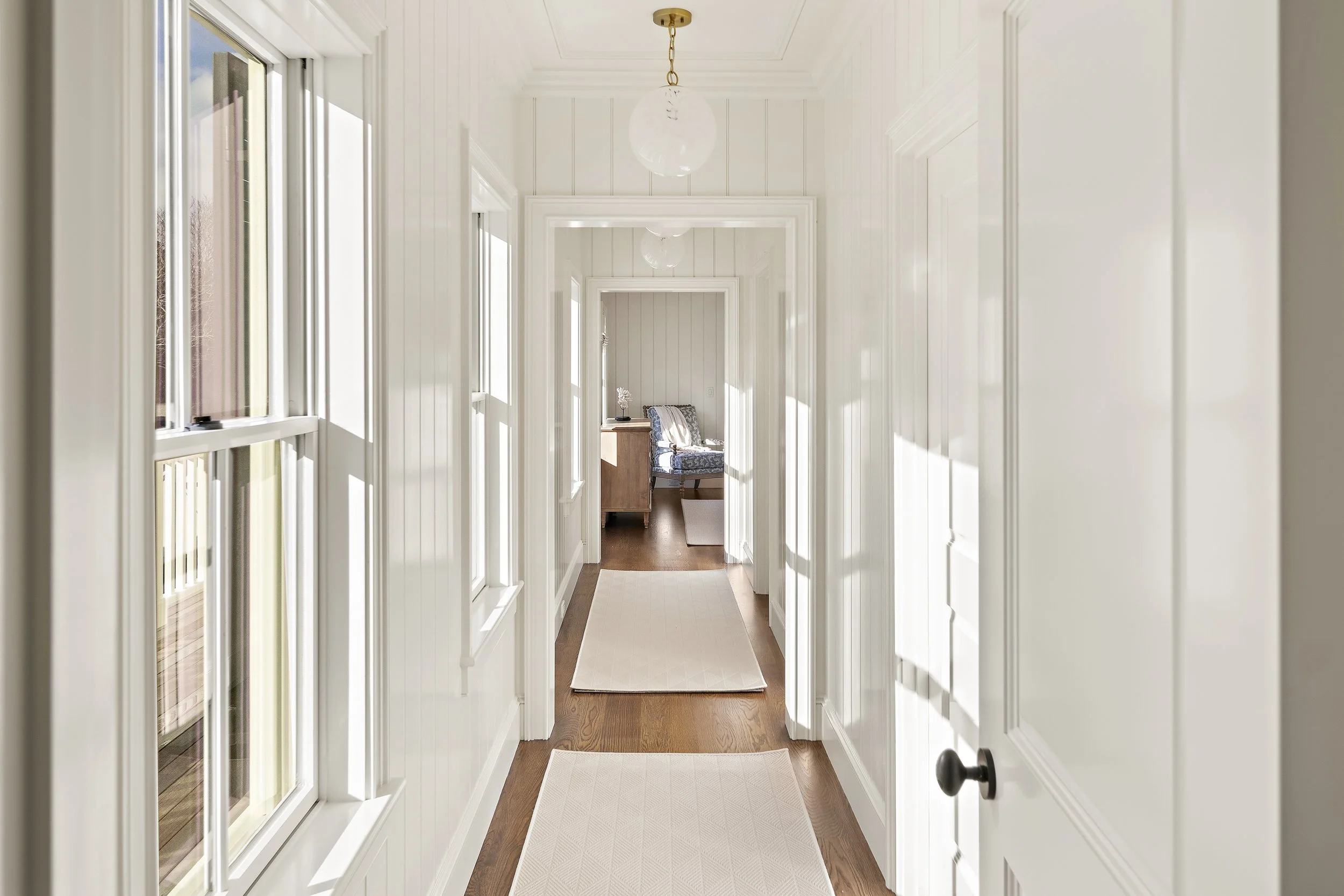 Bright hallway with white paneled walls, hardwood floor, and two white rugs. Sunlight streams in through large windows on the left, leading to a room with a chair, a small table, and a lamp.