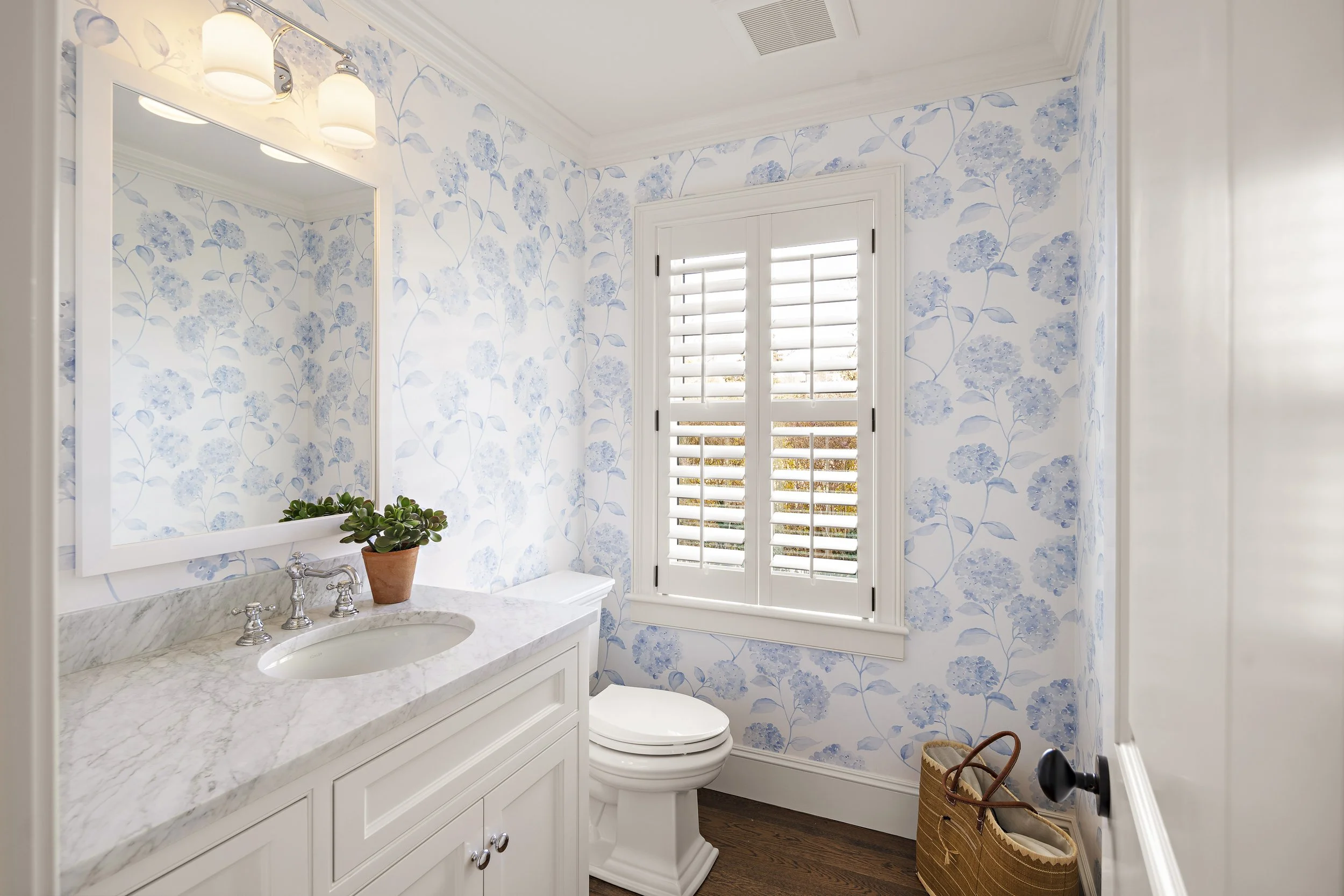 A small bathroom with a floral blue and white wallpaper, a white marble countertop with a sink, a mirror, and a window with white shutters. There is a potted plant on the counter and a woven basket on the floor.