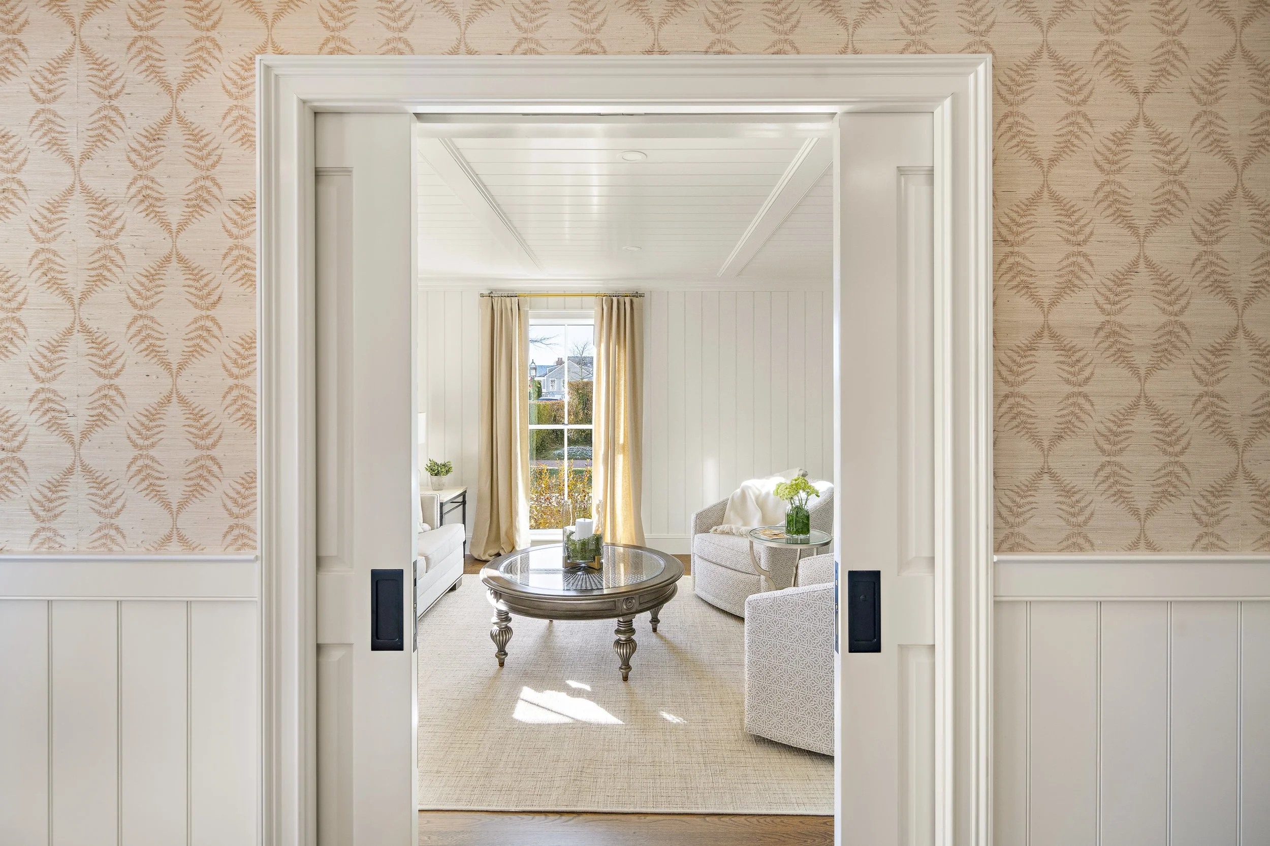 View through a doorway into a bright, cozy living room with a window, beige curtains, white walls, and activity decor.