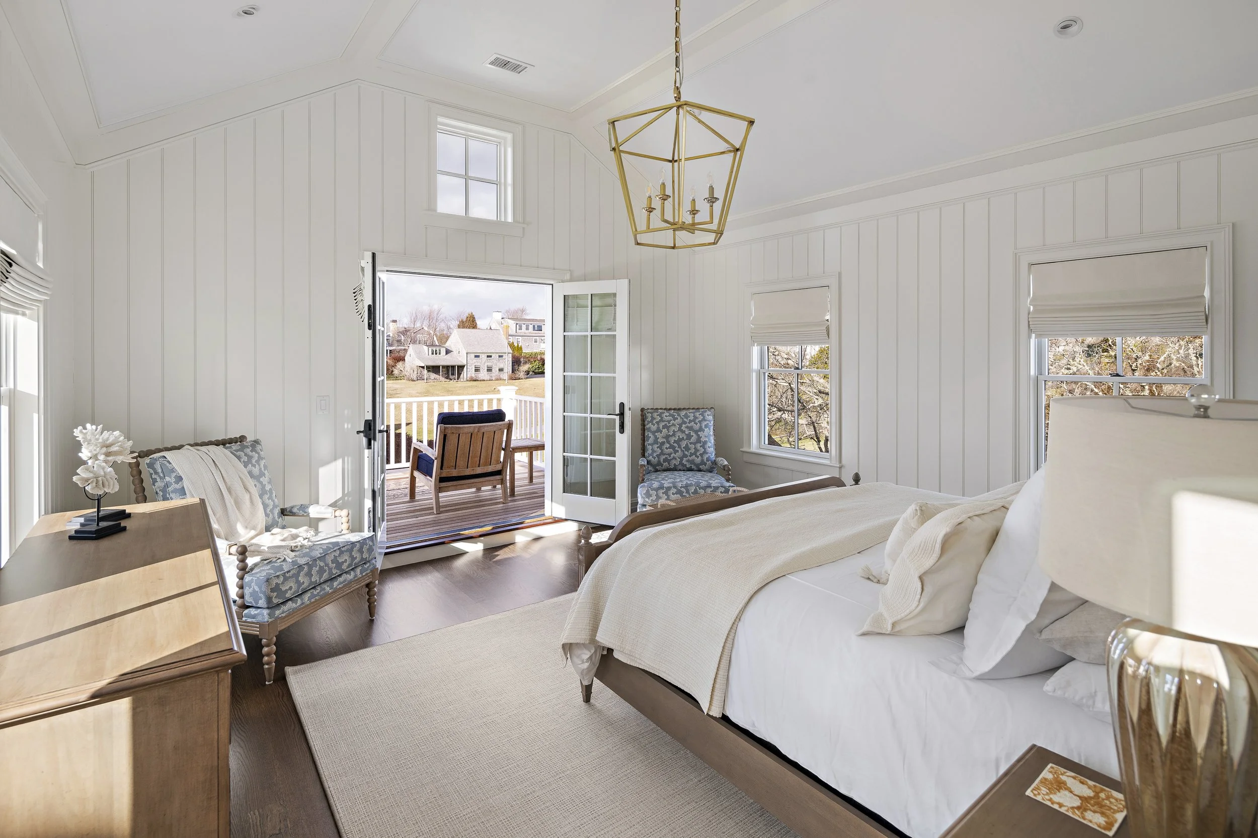 A bright bedroom with white walls and ceiling, featuring large windows and a door opening to a deck with outdoor furniture. The bed has white bedding and pillows, with a nightstand and decor. Two chairs are near the windows, and a chandelier hangs fr