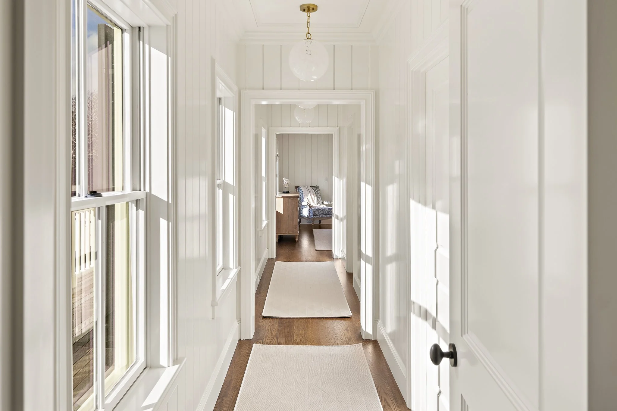 Bright, narrow hallway with white beadboard paneling and large windows on the left side, leading to a room with a wooden floor, a chair with a patterned cushion, and a small side table with a white decorative object.