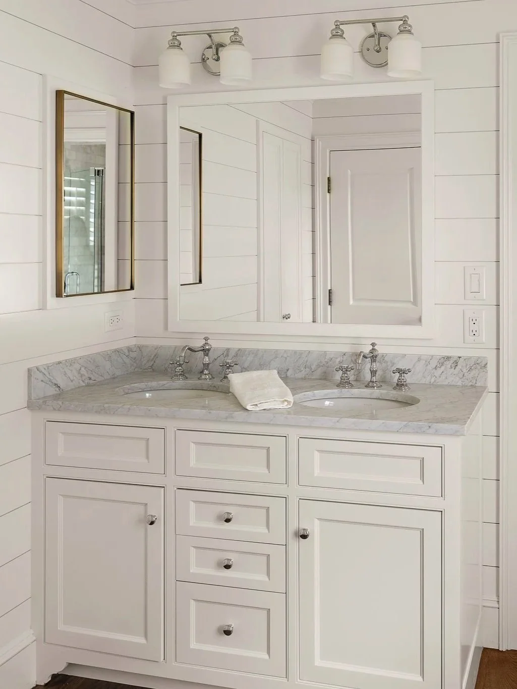 White bathroom vanity with marble countertop, dual sinks, and chrome fixtures, large mirror, wall-mounted light fixture, and shiplap walls.
