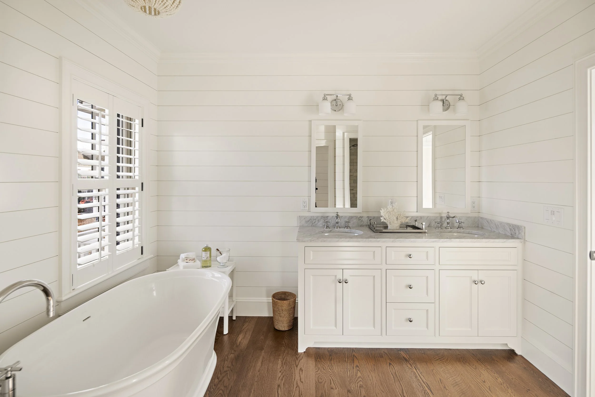 Bright white bathroom with double vanity, marble countertop, mirrors, wall-mounted light fixtures, shiplap walls, a bathtub, and a window with plantation shutters.
