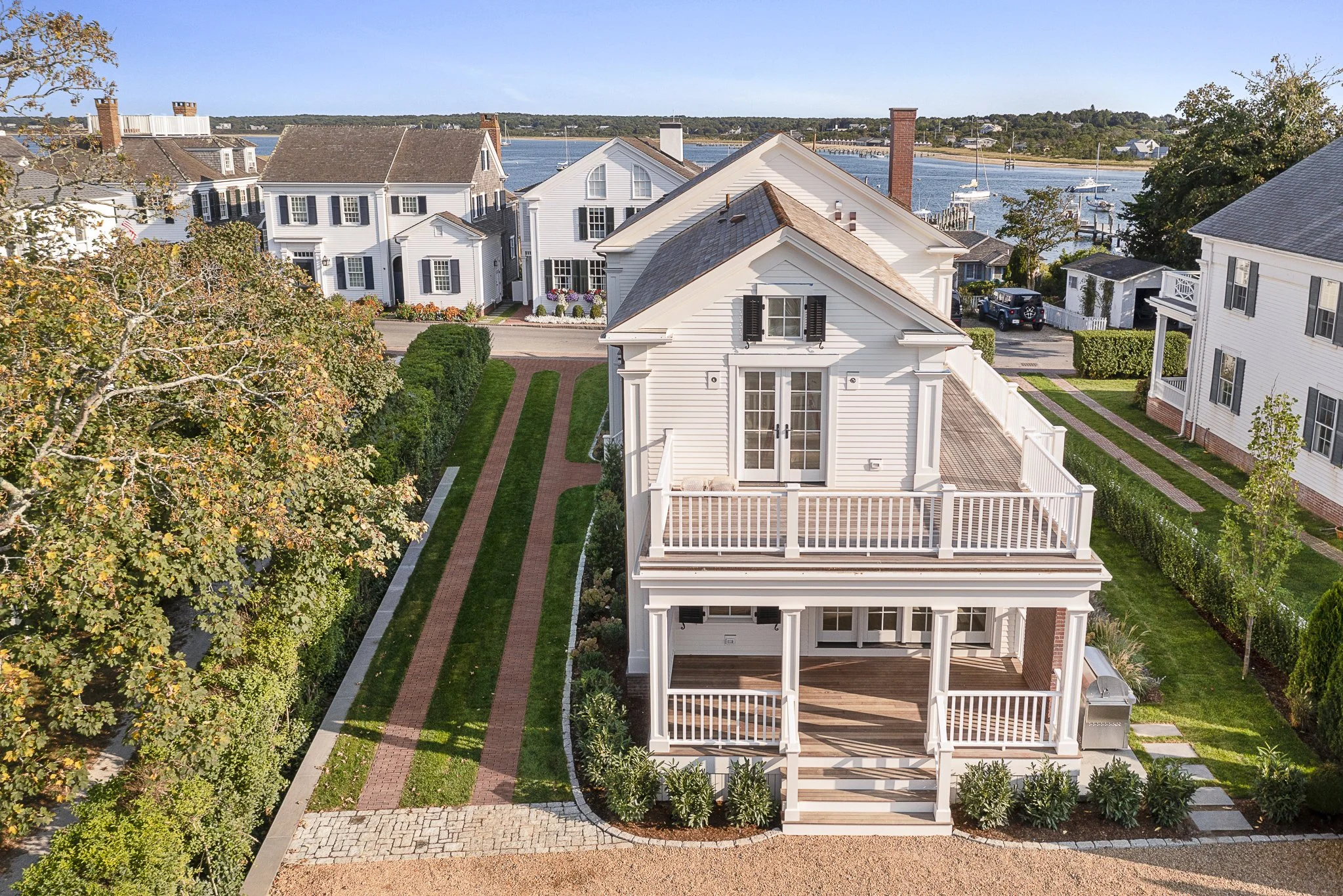 Aerial view of white houses with black shutters near a body of water with sailboats, surrounded by green trees and landscaping.