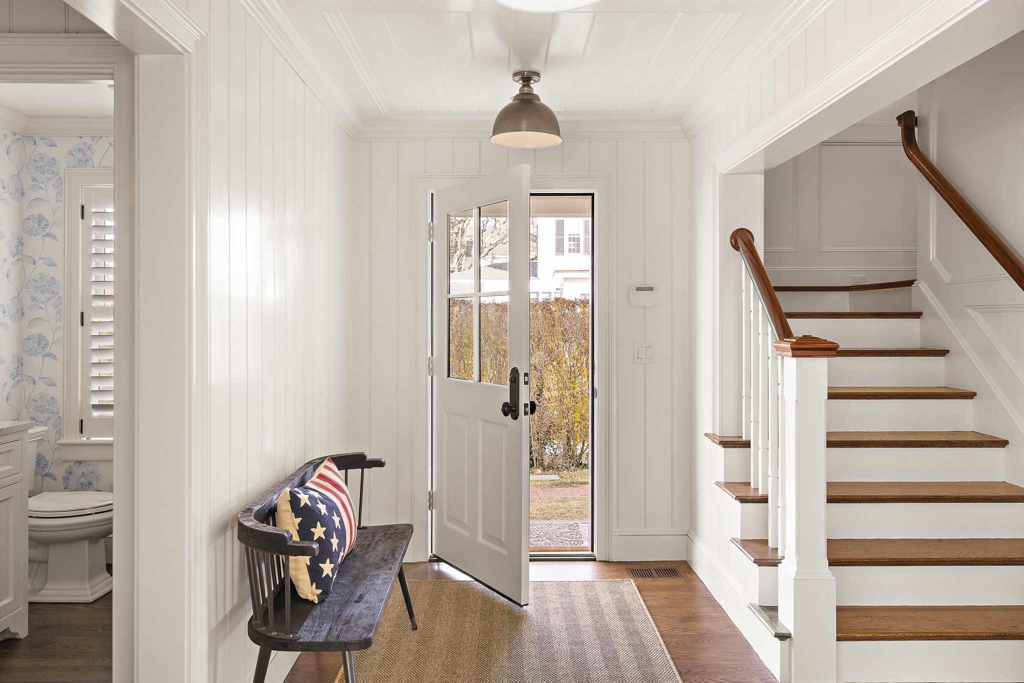 Entryway with front door open to exterior, wooden stairs with white railings, black bench with star-patterned cushion, and a bench nearby. The interior features white paneled walls, hardwood flooring, and a ceiling-light fixture. Peeks of wallpaper with blue floral pattern and a toilet are visible on the left.