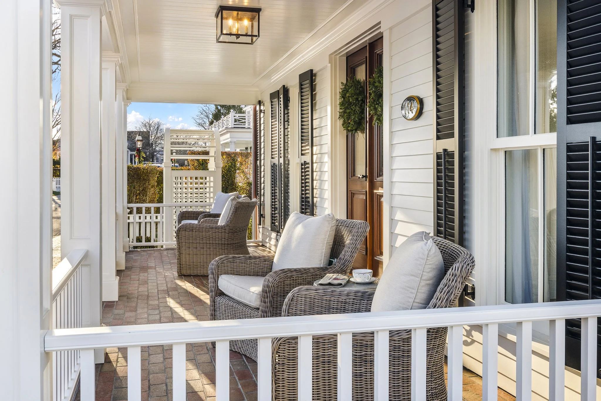 Front porch of a house with white railings, wicker chairs with cushions, and a brick floor, decorated with holiday greenery and ornaments, against a background of trees and other houses under a blue sky.
