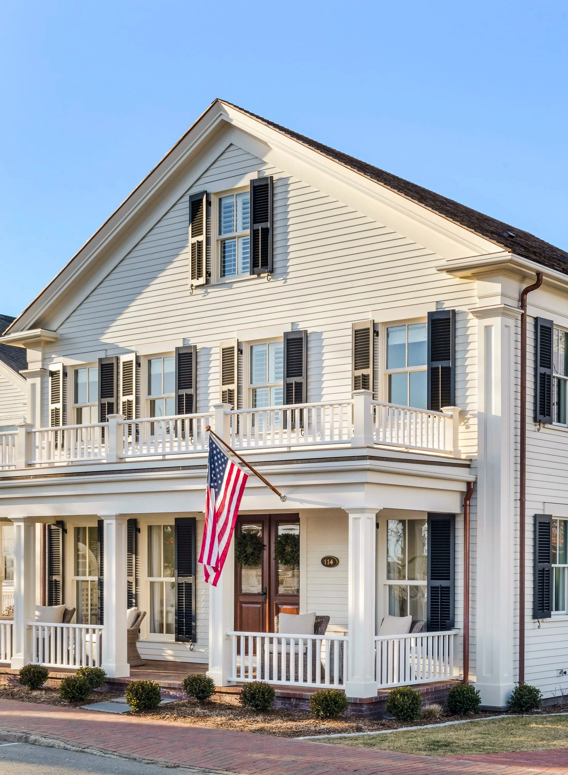 Large white house with black window shutters, front porch with white railing, American flag, and outdoor furniture, under a clear blue sky.