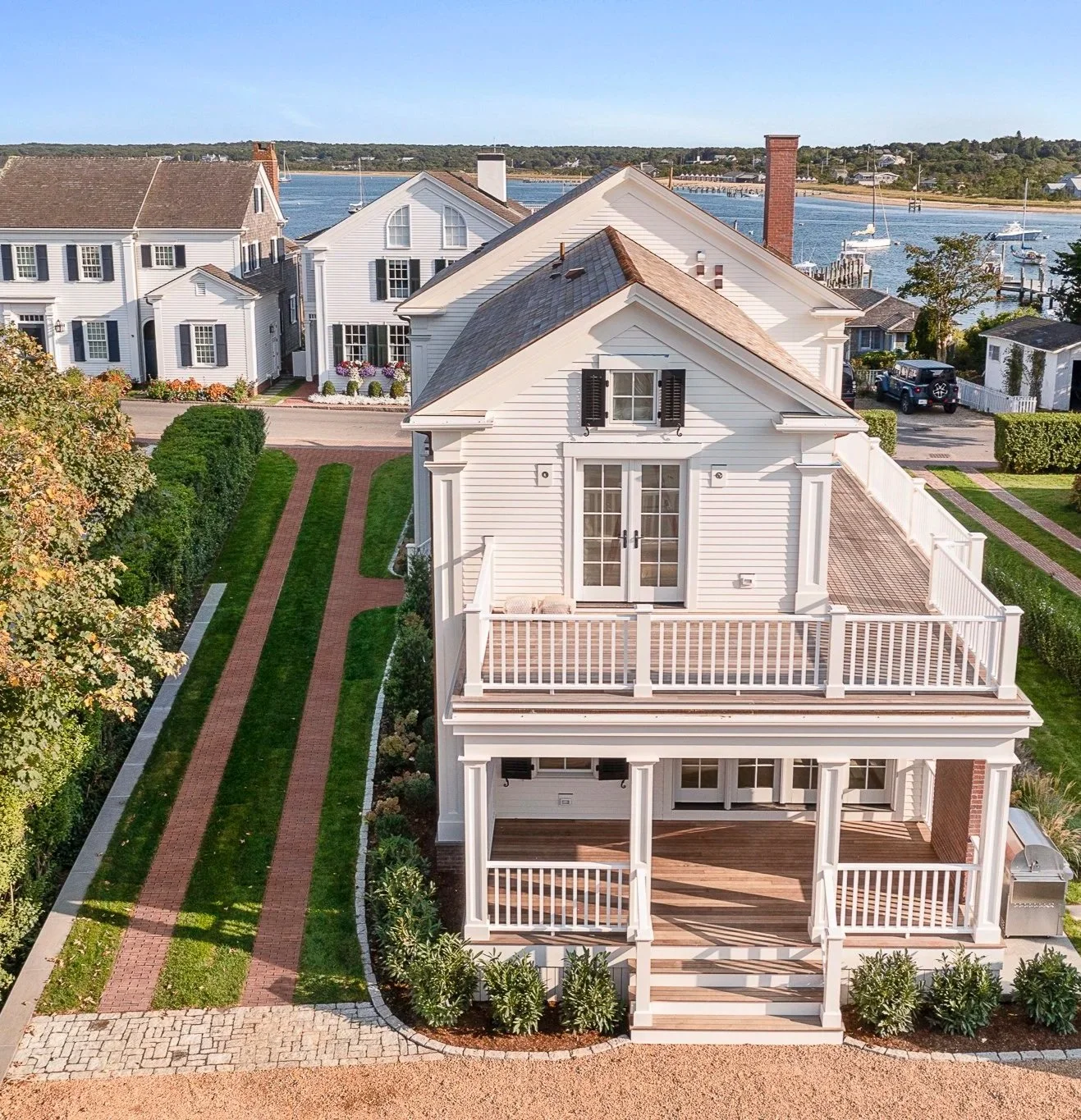 Aerial view of a white, multi-story house with a waterfront in the background, including boats docked at the water's edge, and a brick pathway leading to the house front. The house has a balcony with white railings and a large deck on the second floor with a porch below.