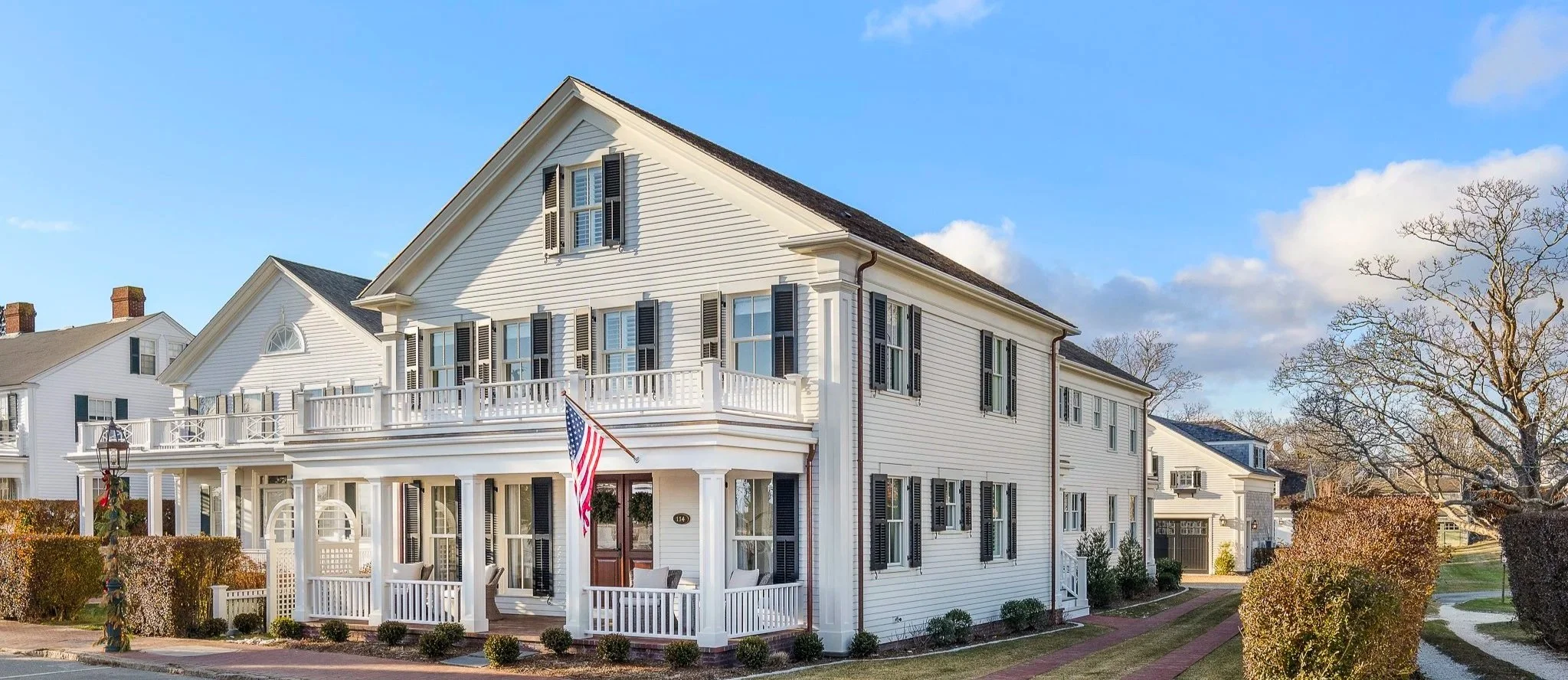 White multi-story house with black shutters, front porch, balcony, and American flag, set in a neighborhood with trees and blue sky.