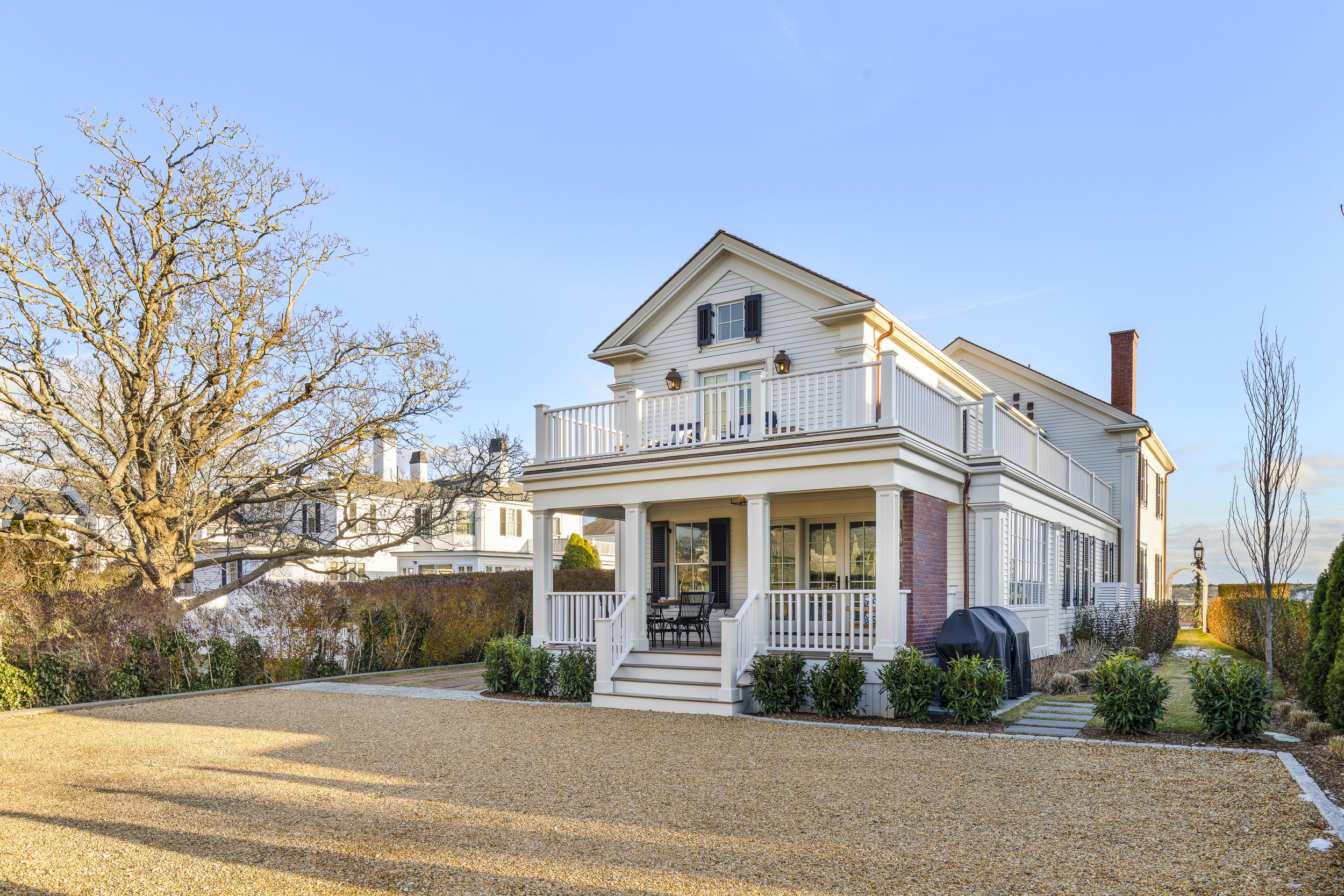 Backyard view of a large white house with black shutters, a brick chimney, and a spacious porch with outdoor furniture, surrounded by leafless trees and a well-maintained garden under a clear blue sky.