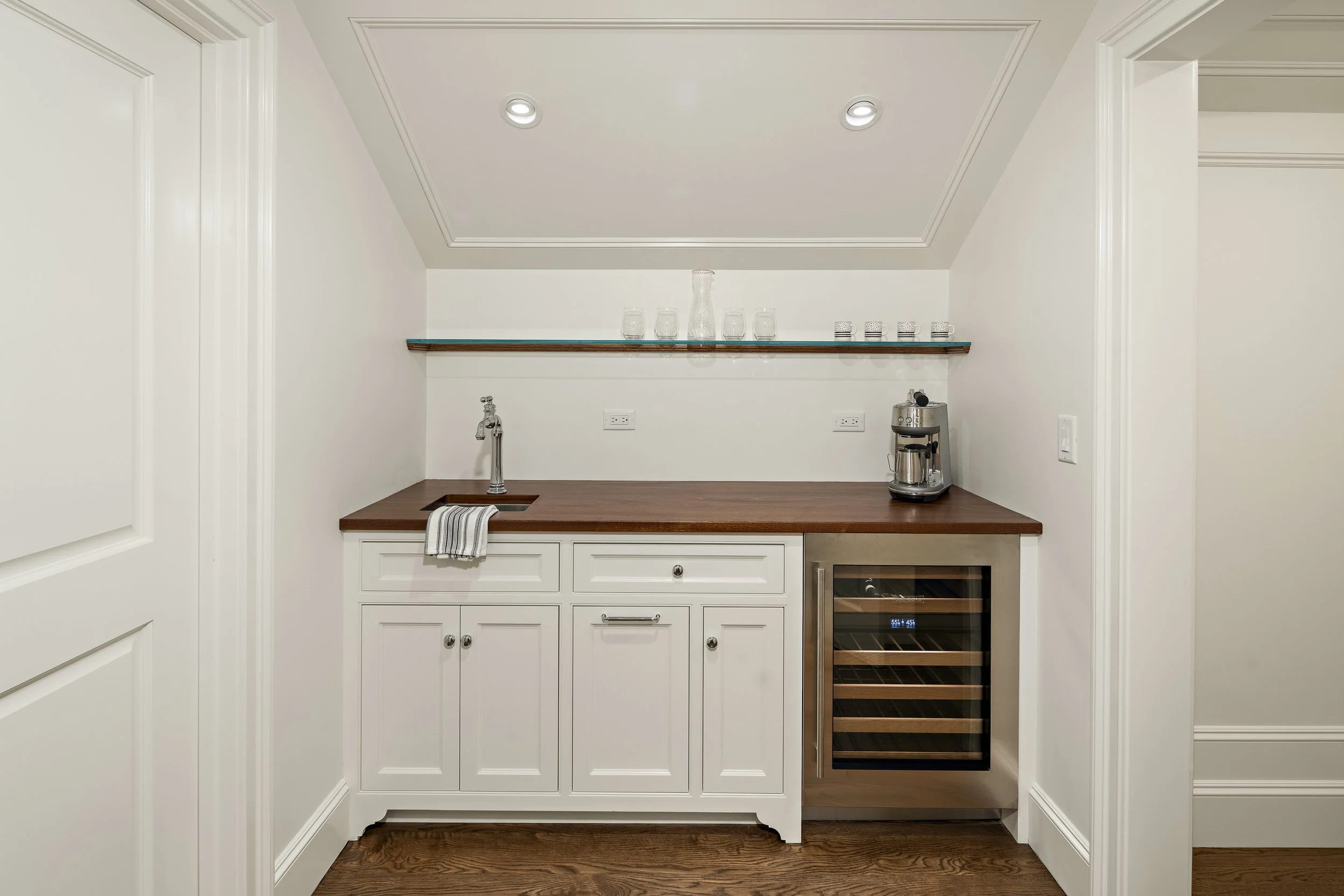 A small kitchenette with white cabinetry, a wooden countertop, and a wine cooler. There are glassware and a decorative glass pitcher or vase on a glass shelf above the counter. The room has recessed lighting and electrical outlets on the wall.
