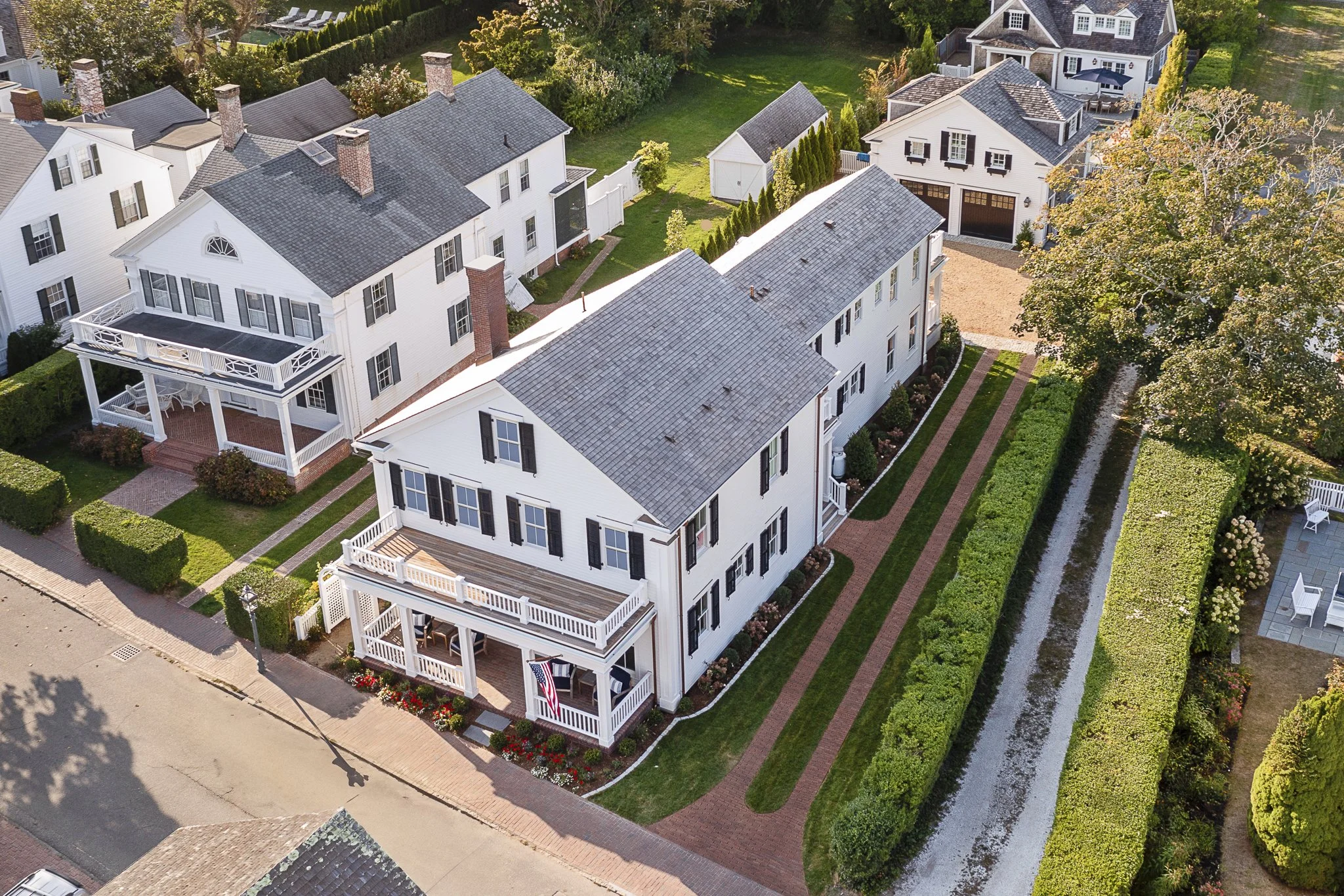 An aerial view of a residential neighborhood featuring multiple houses with gray shingle roofs, white exterior walls, and black shutters. Some houses have front porches and balconies. The houses are surrounded by well-maintained lawns, gardens, and trees. There is a narrow canal or waterway running along the right side of the image, with a hedge and trees lining it. A brick sidewalk runs in front of the houses with parking spaces and a flagpole visible.