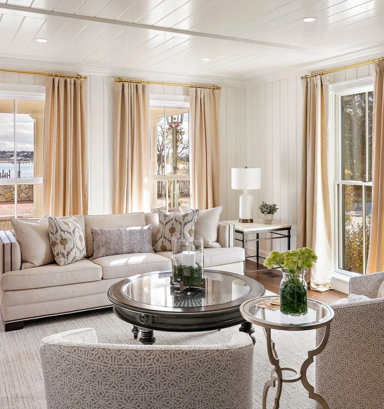 Bright living room with beige curtains, white paneled walls, and large windows showing an outdoor view. There is a white sofa with patterned and solid throw pillows, a glass coffee table with a candle centerpiece, a side table with a white lamp, potted plant, and a vase of green flowers, and a patterned armchair.