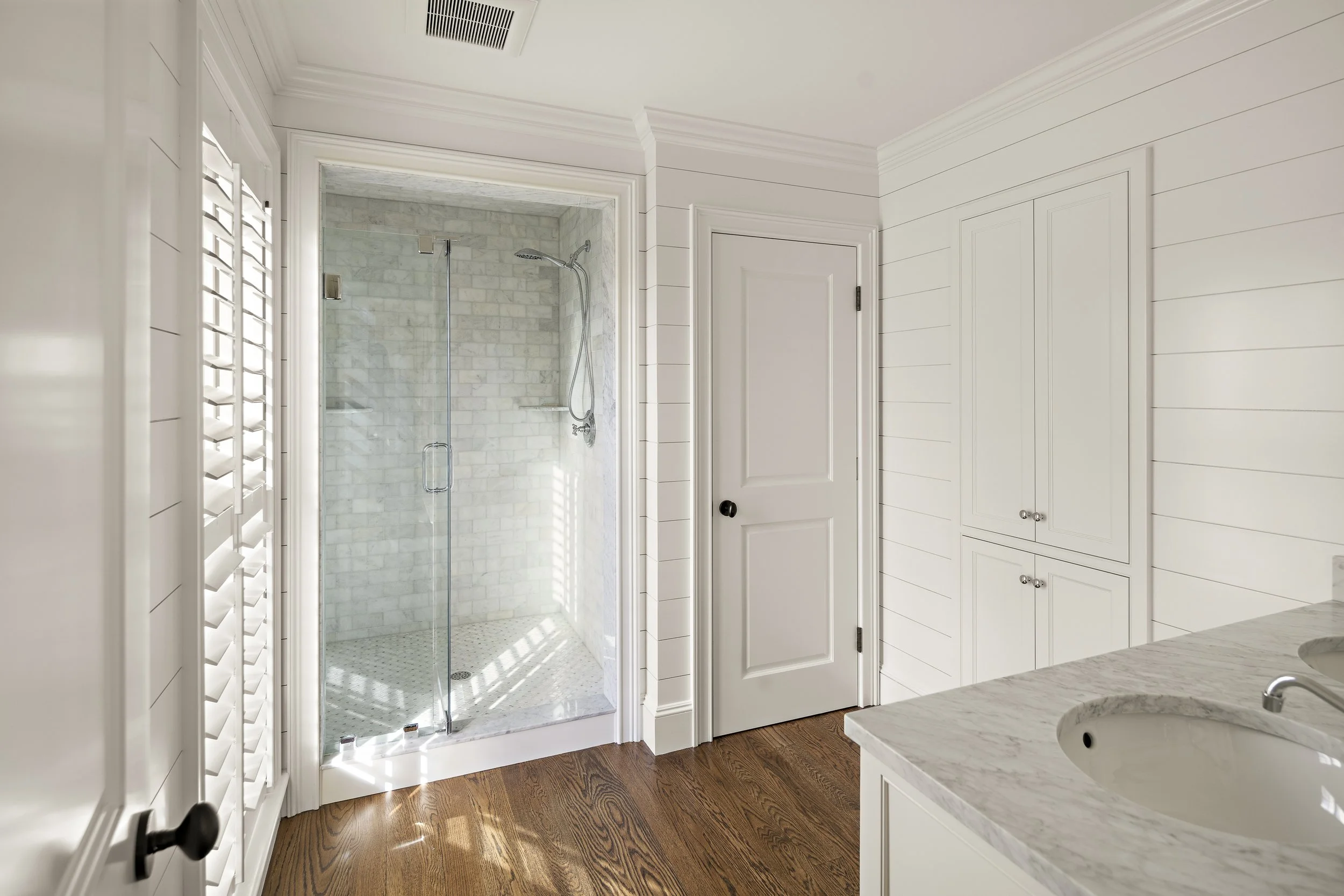 A bathroom with white shiplap walls, a glass walk-in shower with marble tiles, a white countertop with a marble top, and wood flooring.