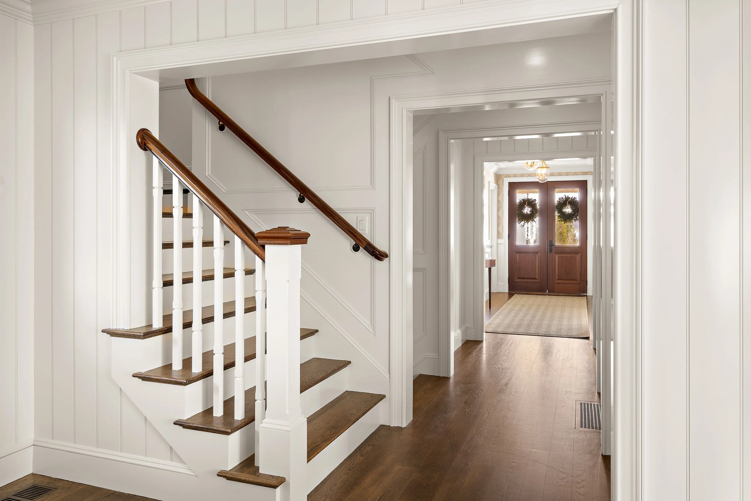 A hallway with white walls and wooden flooring, featuring a staircase with white balusters and a wooden handrail, and a dark wooden front door decorated with two wreaths.