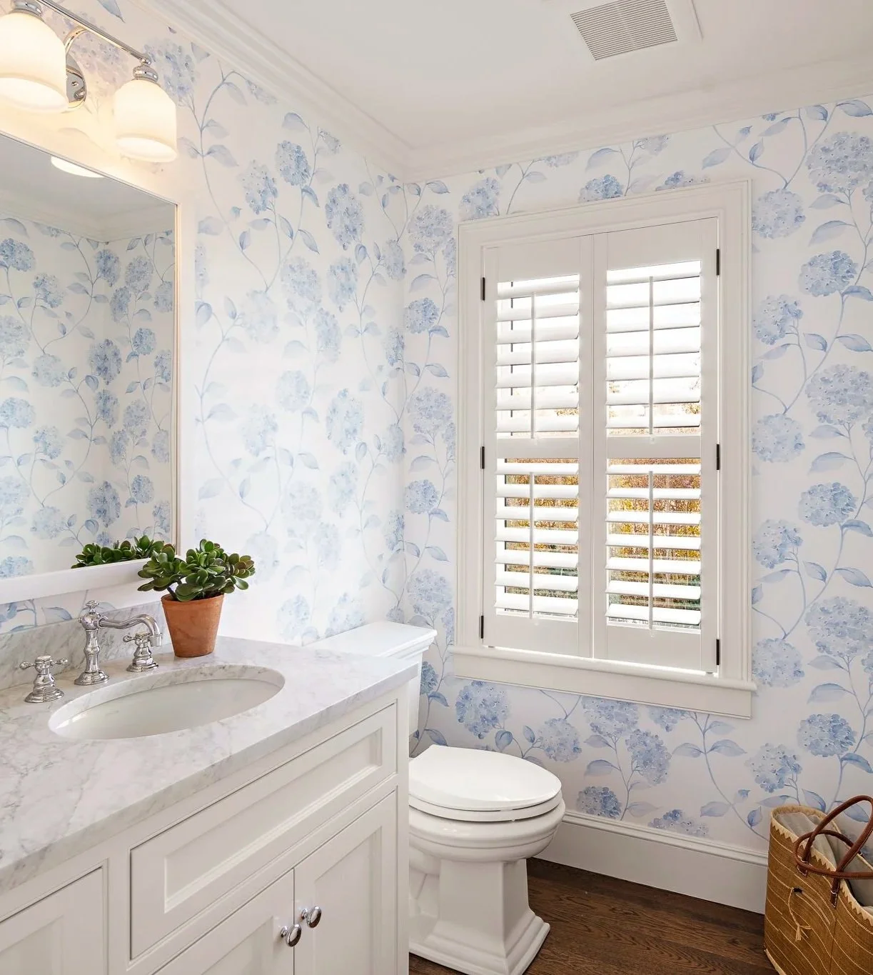 A bright bathroom with floral wallpaper, a white vanity with a marble countertop, a potted plant, a toilet, a large window with shutters, and a wicker basket.