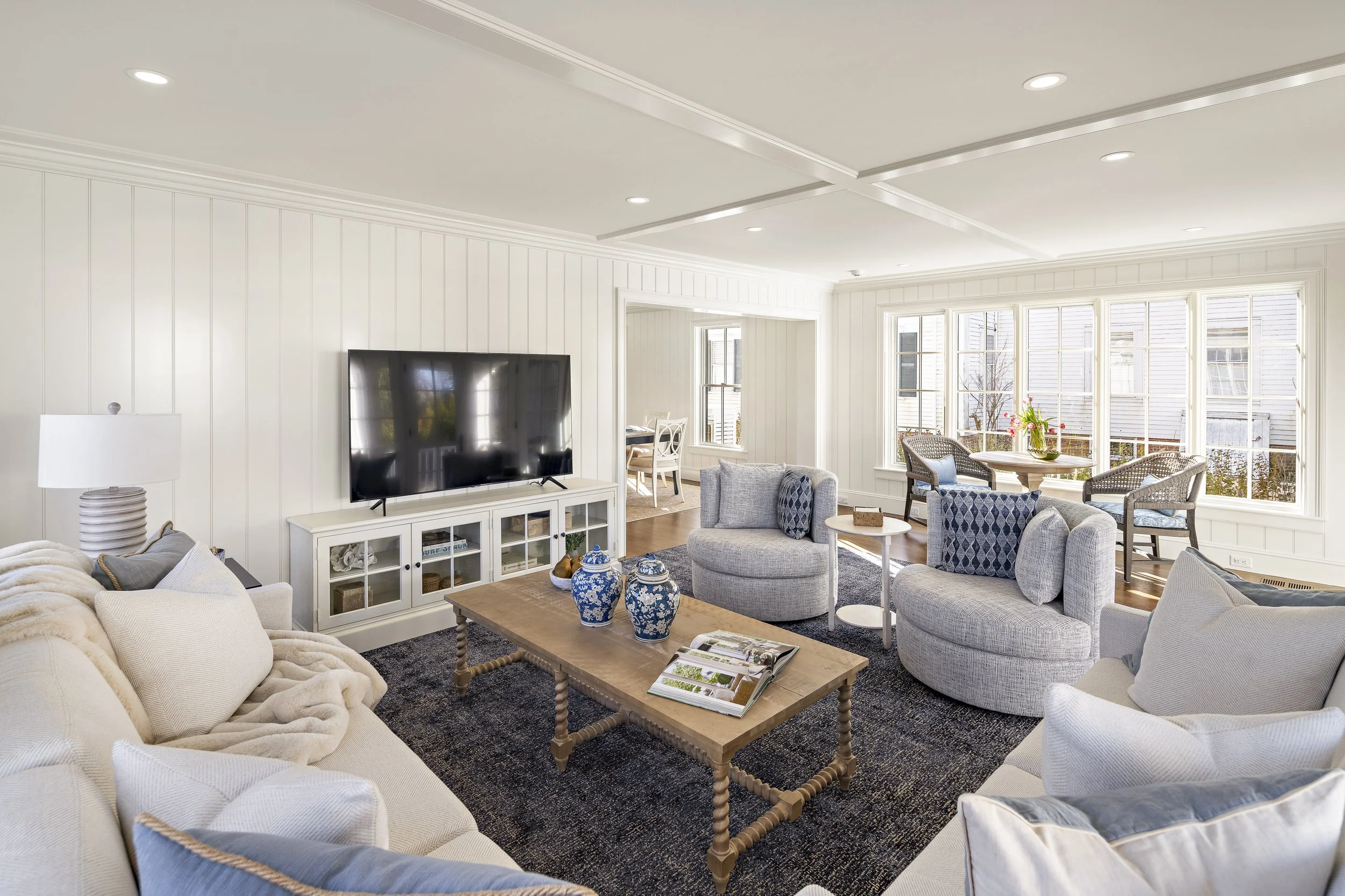 A spacious living room with white walls and large windows allowing natural light. There are multiple sofas, a coffee table with decorative blue and white vases, and a TV on a white cabinet. The room has a cozy and bright ambiance with a mix of modern