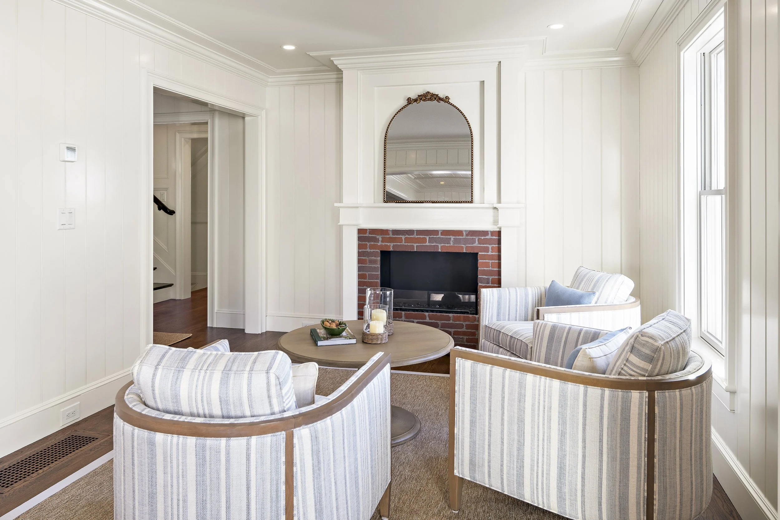 Living room with white paneled walls, a brick fireplace with a mirror above it, wooden armchairs with striped cushions, a round wooden table with candles and a plant, and large windows letting in natural light.