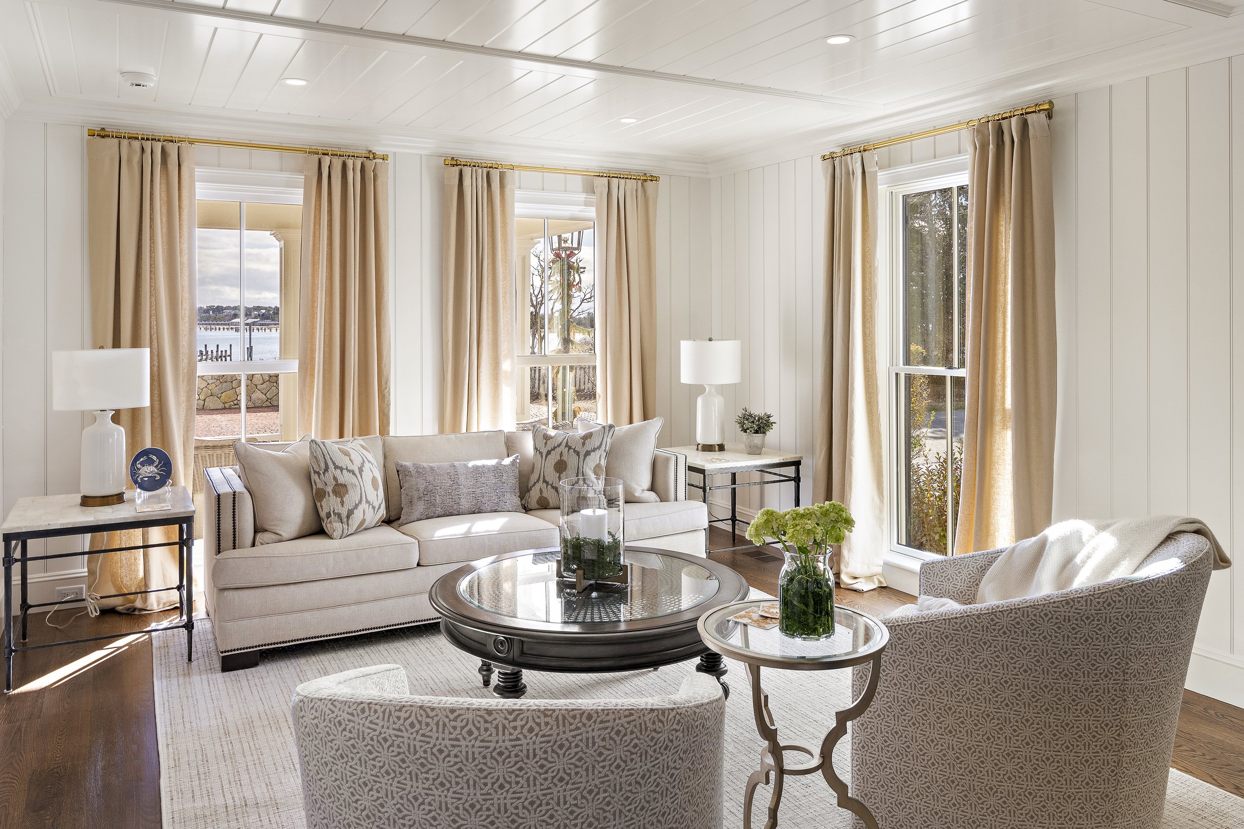 Bright living room with white paneled walls and ceiling, large windows with beige curtains, beige sofa with decorative pillows, glass coffee table with floral arrangements, and a side table with lamps.