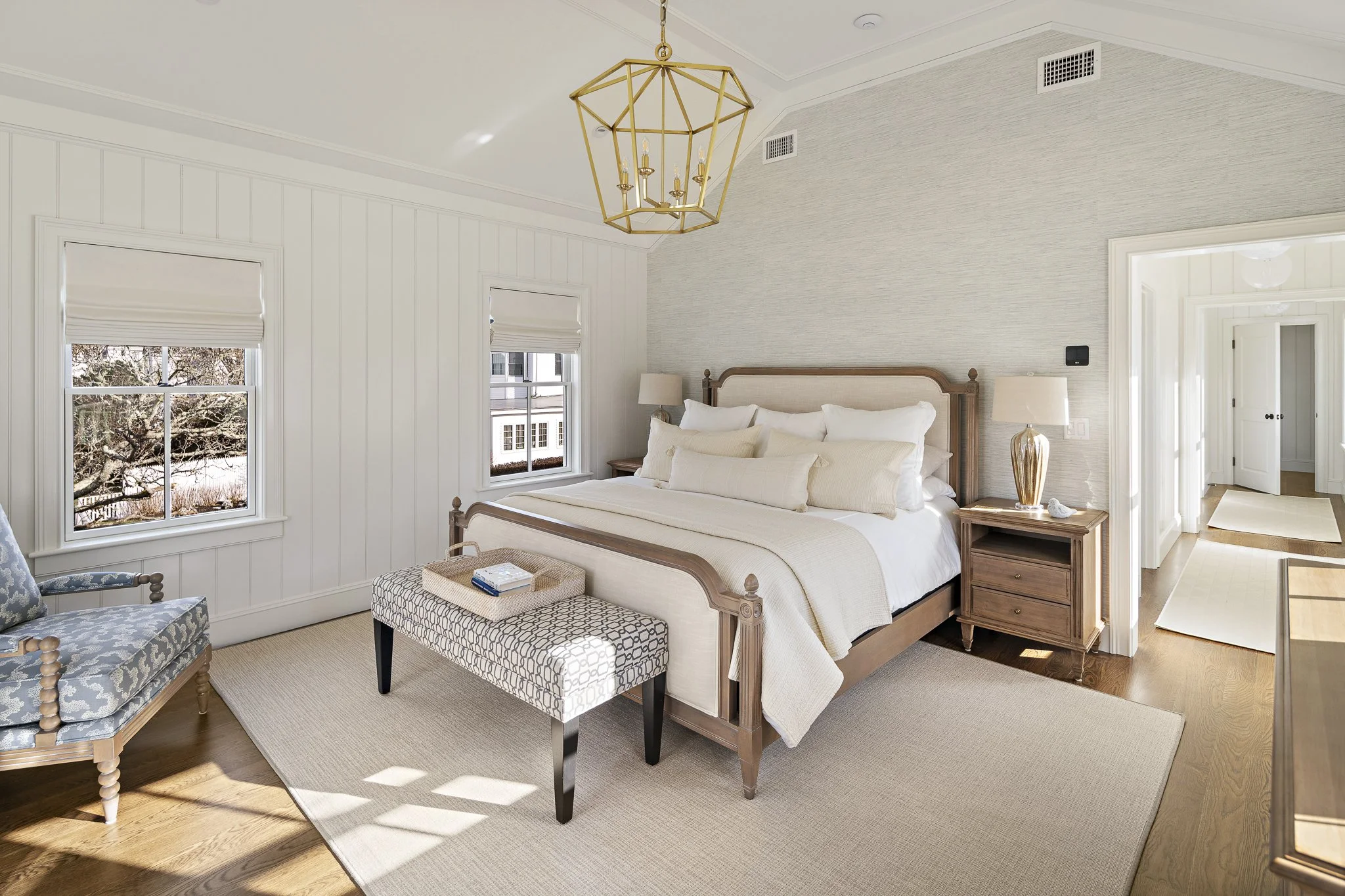 A bedroom with a wooden bed frame, white bedding, a pair of matching nightstands with lamps, a patterned ottoman, two windows with blinds, a chandelier, and a doorway leading to another room.