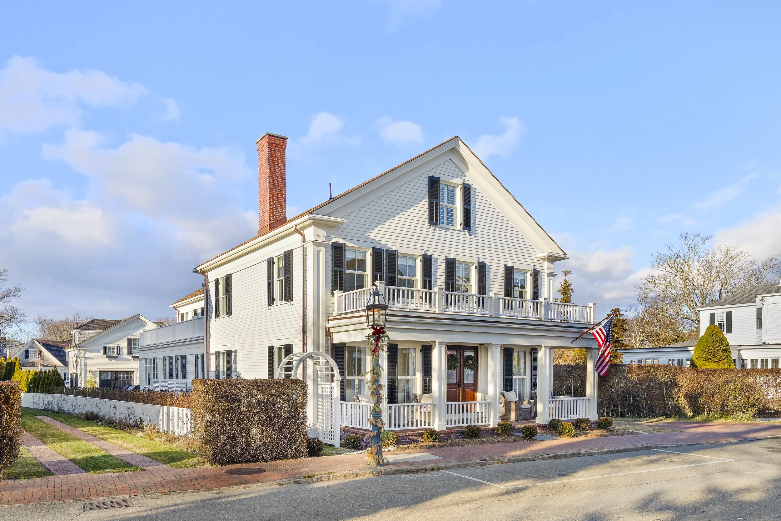 White multi-story house with black shutters, a brick chimney, and a front porch with outdoor furniture, under a partly cloudy blue sky. Decorated for the holidays with an American flag, Christmas wreath, and ribbon on the porch.