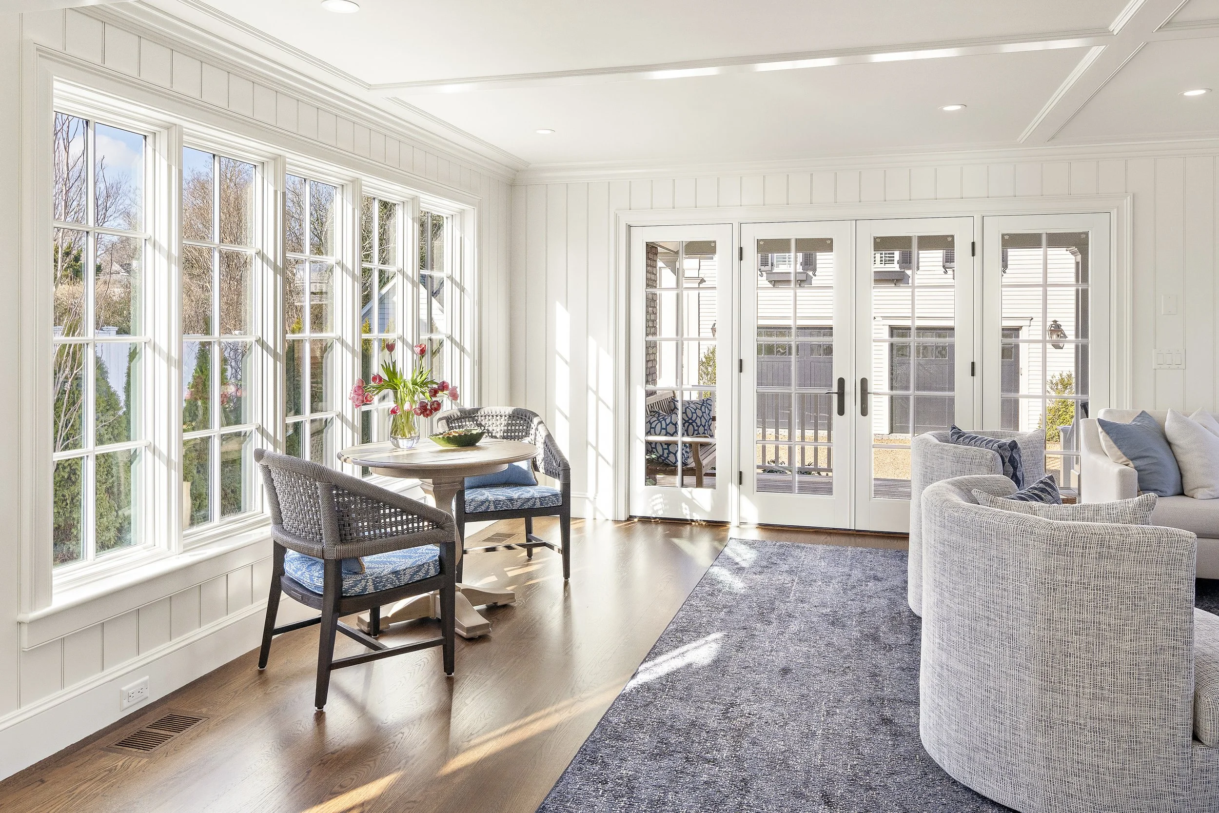 A bright sunlit living room with large windows, a wooden dining table with chairs and a vase of pink flowers, and a cozy seating area with a light-colored sofa and chairs, hardwood floors, and a dark area rug.