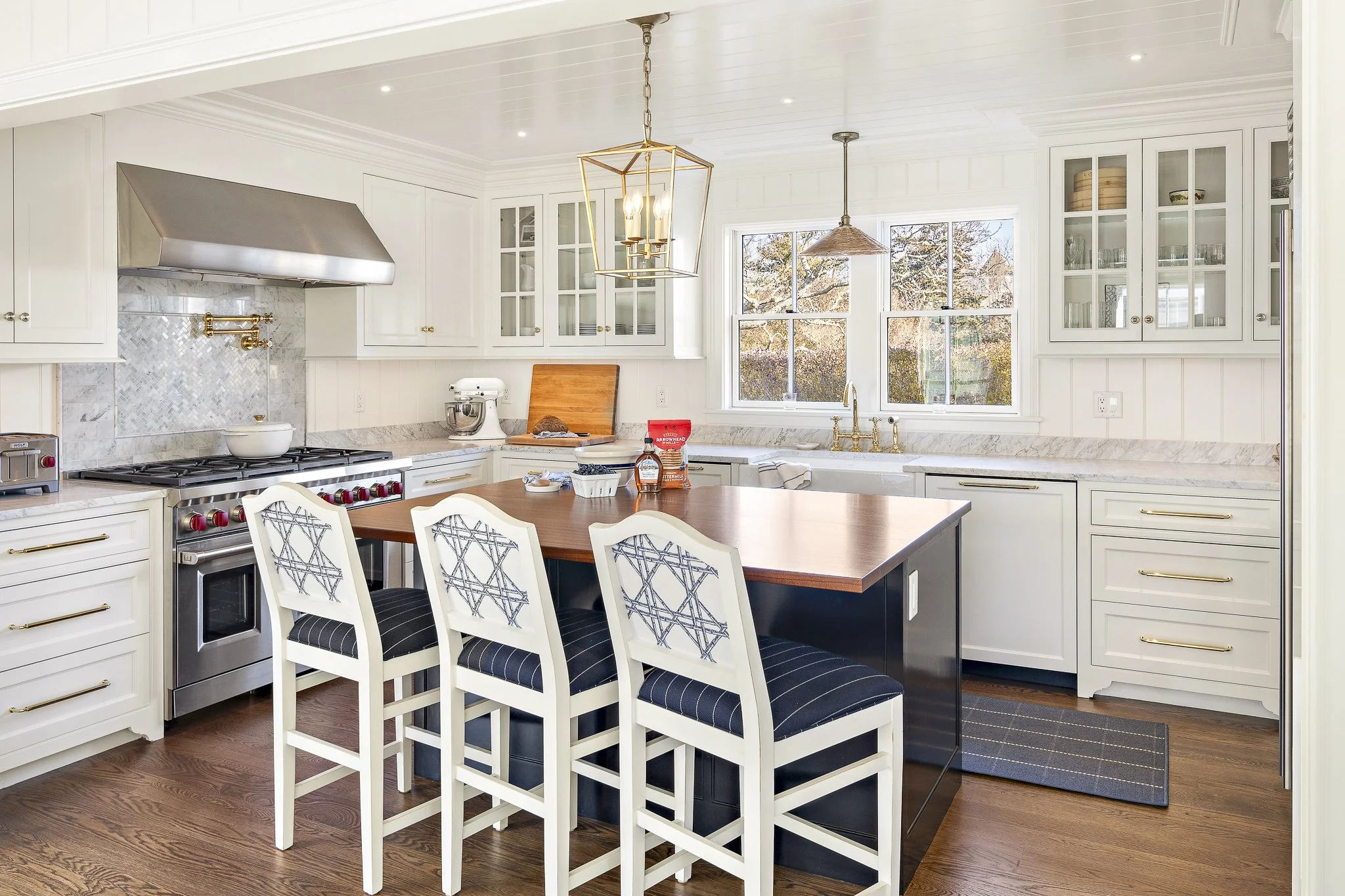 Bright, modern kitchen with white cabinets, marble backsplash, and a large window overlooking trees. A wooden dining table with white and blue chairs is in the foreground. Kitchen appliances include a stainless steel stove, oven, and a stand mixer. Decor includes a gold geometric chandelier and a woven pendant light.