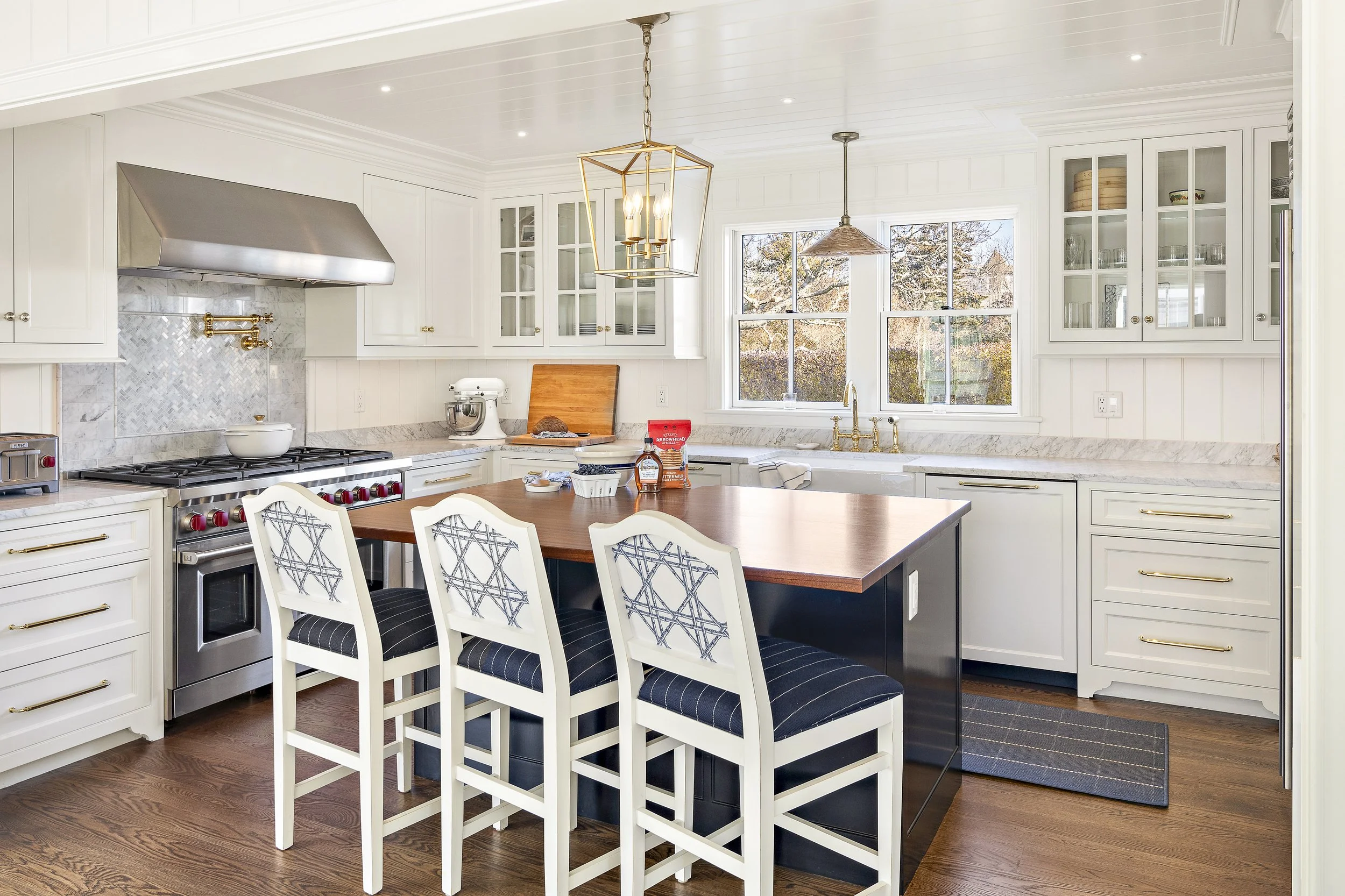 Bright kitchen with white cabinets, a marble backsplash, and a large window with a view of trees outside. Features include a stainless steel range hood, a white stand mixer, a cutting board, and a wooden dining table with six chairs. Gold accents are