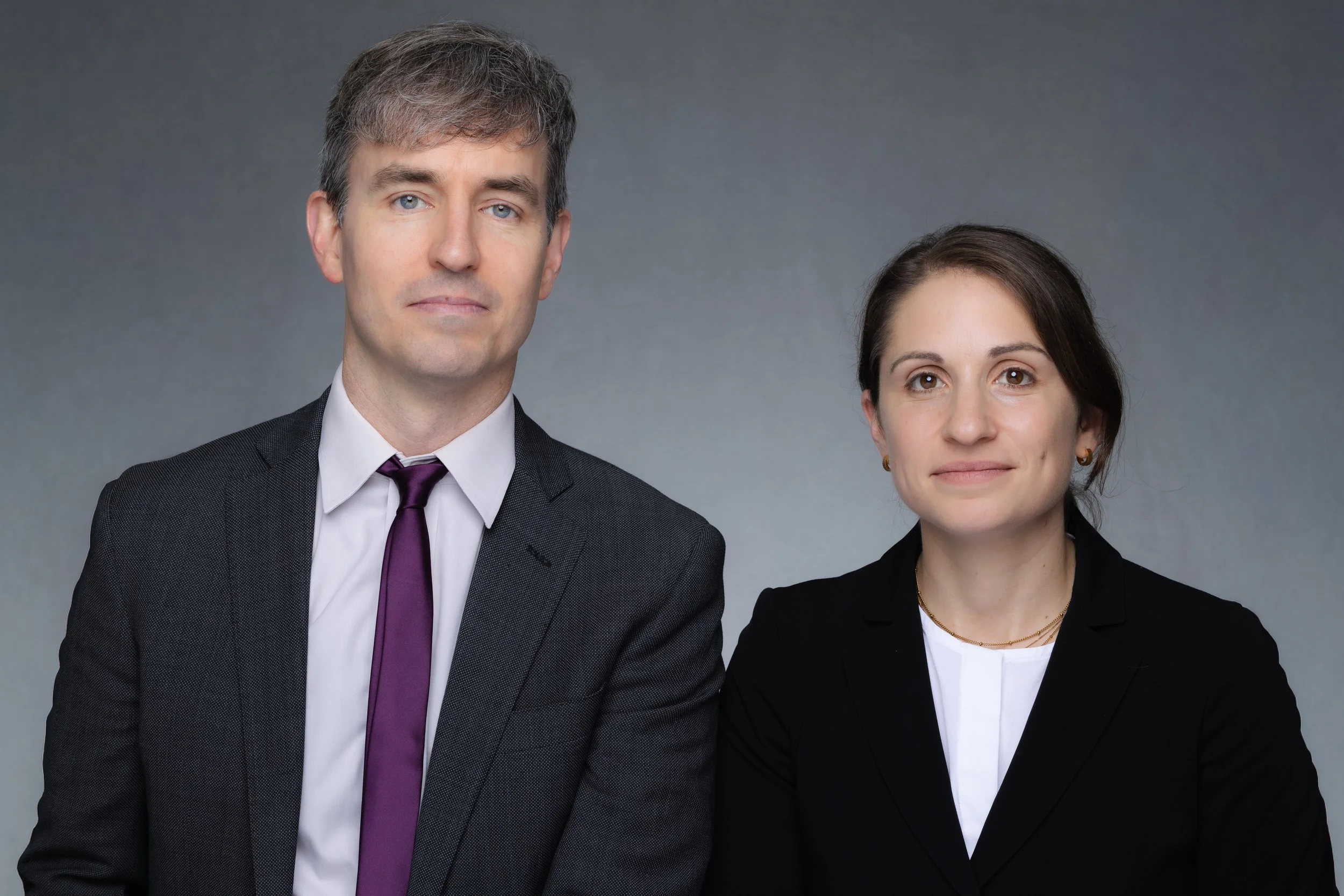 A man and a woman in formal business attire standing against a plain gray background.