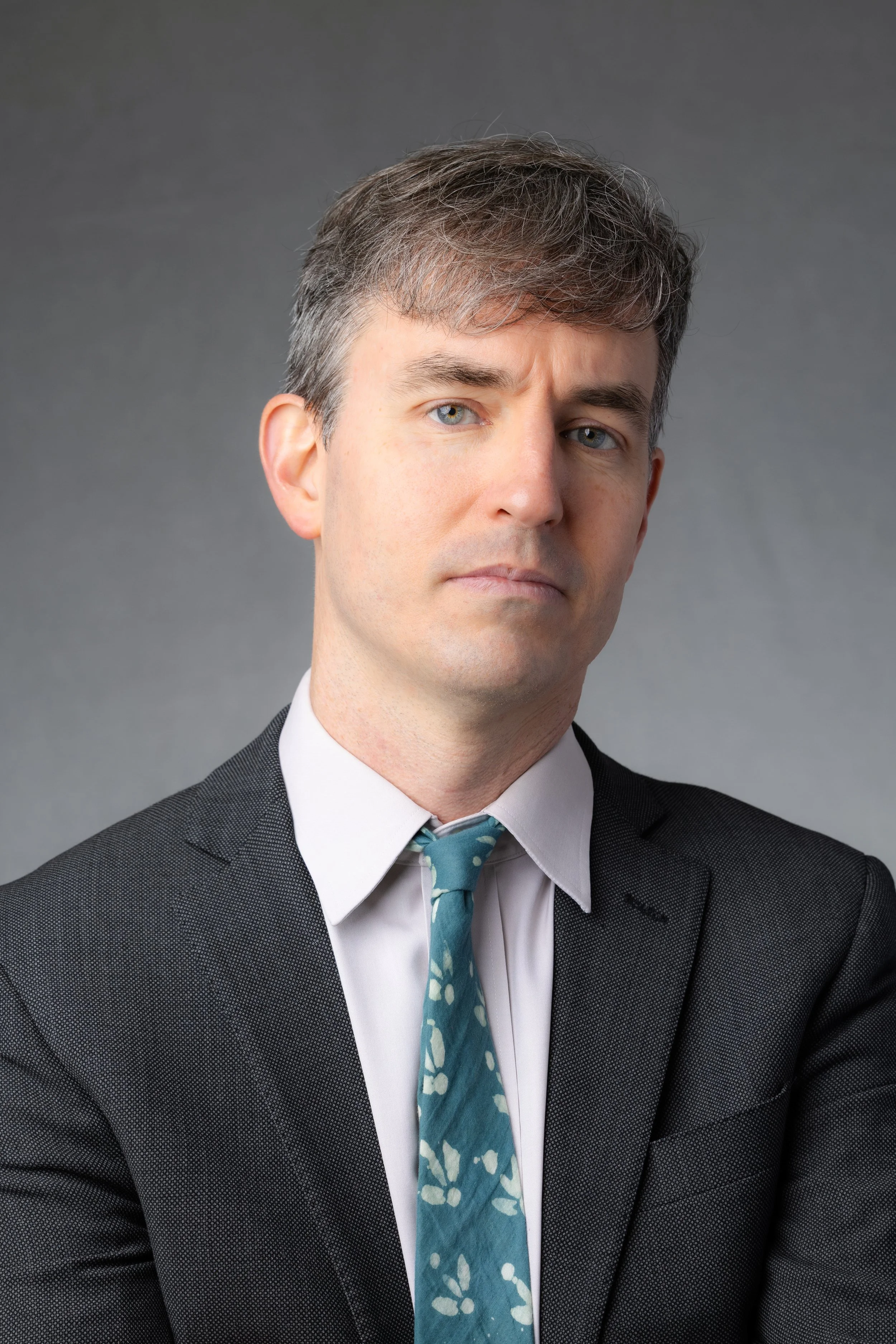 A man in a dark gray suit with a white shirt and a turquoise tie with white floral pattern, posing against a gray background.