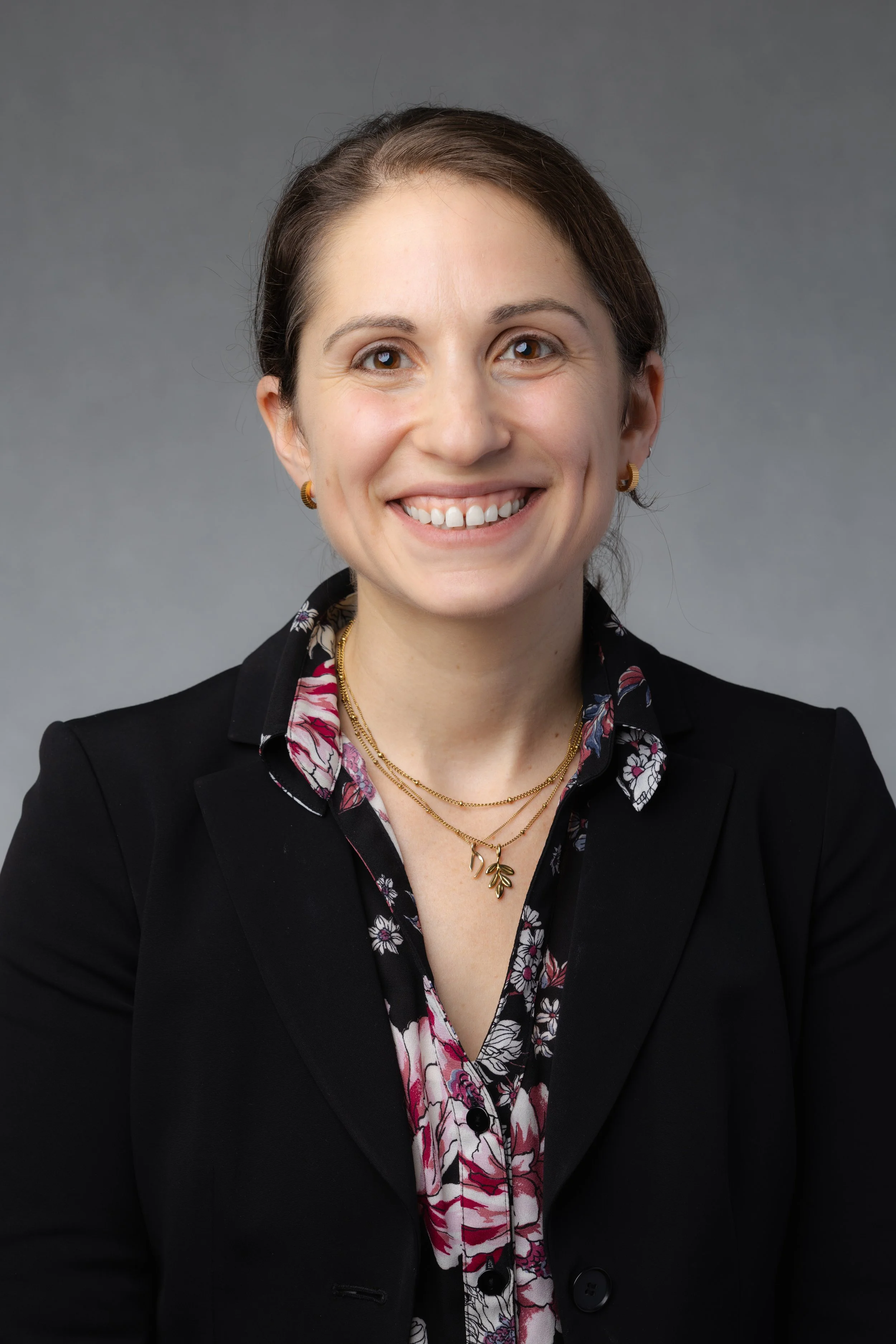 A woman smiling, wearing a black blazer and a floral blouse, with gold jewelry, in front of a gray background.